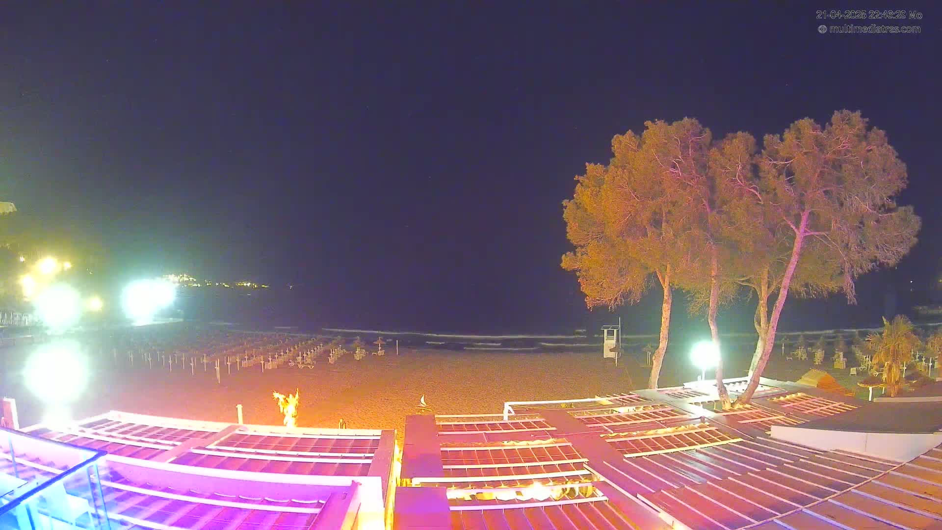 A nighttime beach scene shows a sandy shore with closed beach umbrellas, a calm sea, and several illuminated structures, including tall pine trees under a dark sky.