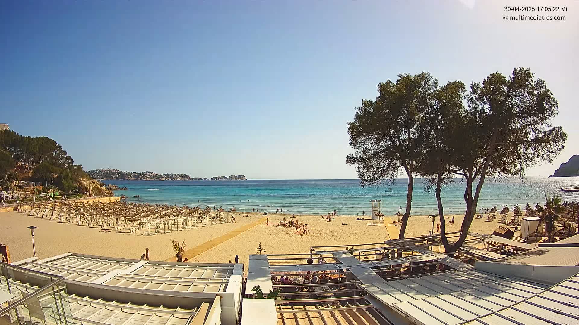 A sandy beach with numerous umbrellas and beachgoers, overlooks a calm, turquoise ocean under a clear blue sky.