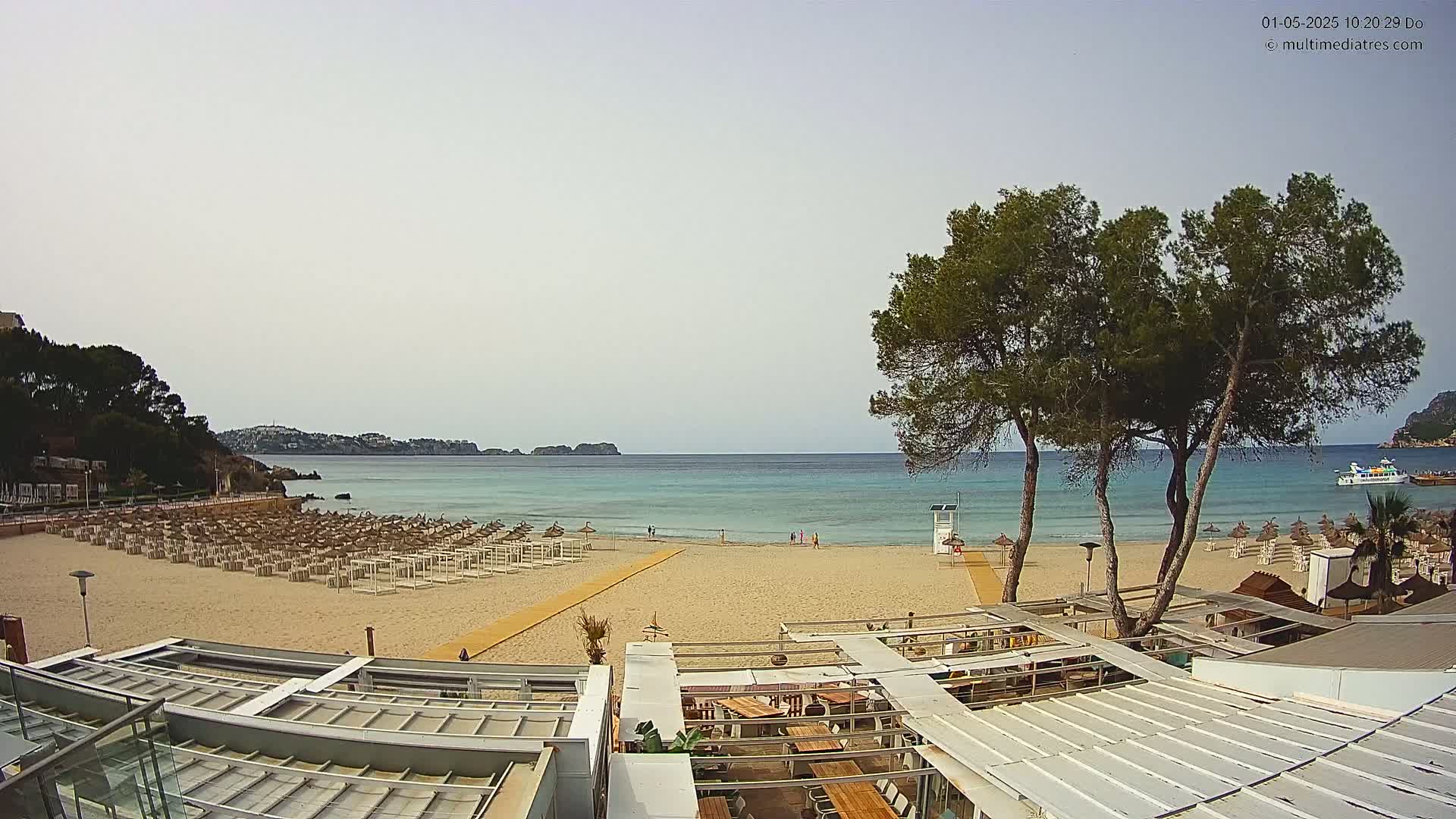 A sandy beach with rows of lounge chairs and umbrellas overlooks a calm, turquoise sea under a mostly clear sky.