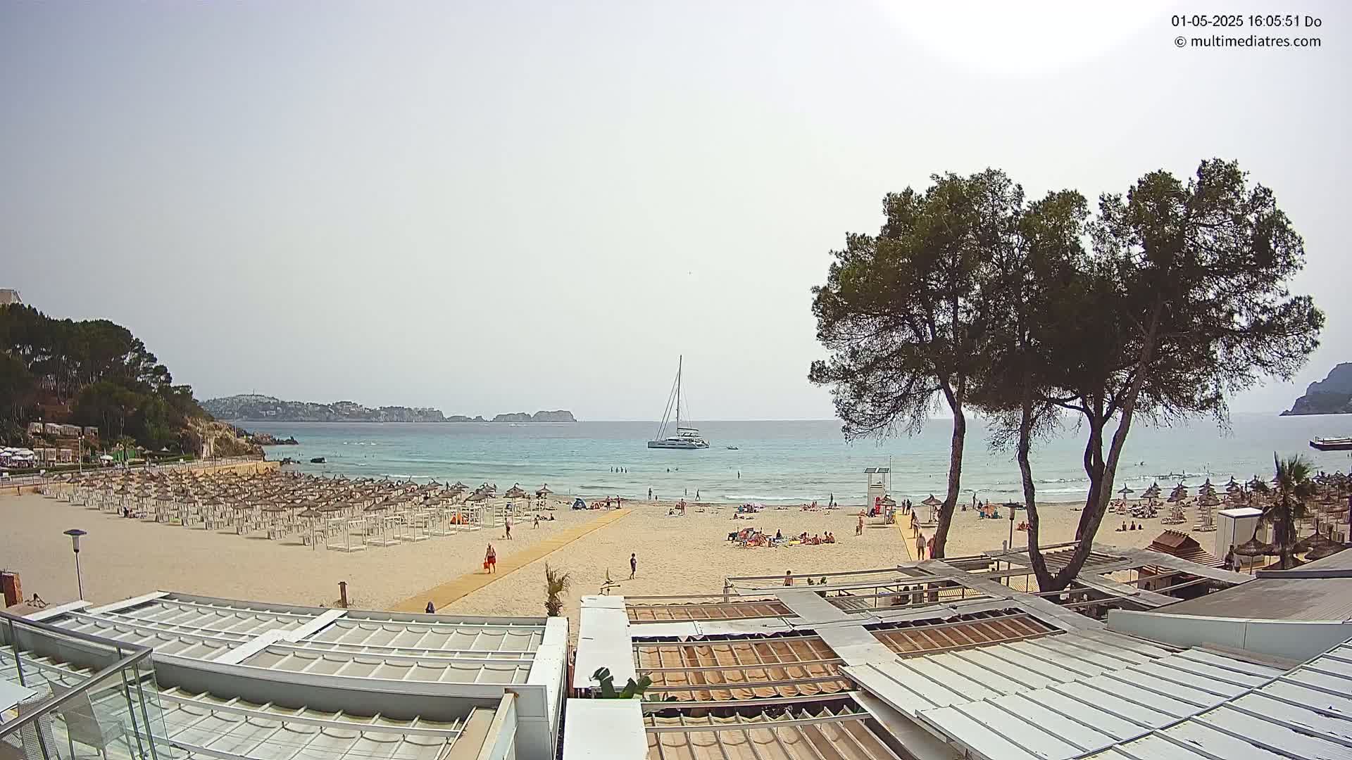 A sandy beach with many umbrellas and sunbathers is partially visible from an elevated vantage point overlooking calm, light-blue water under a hazy sky; a sailboat is anchored offshore, and a few trees frame the view.