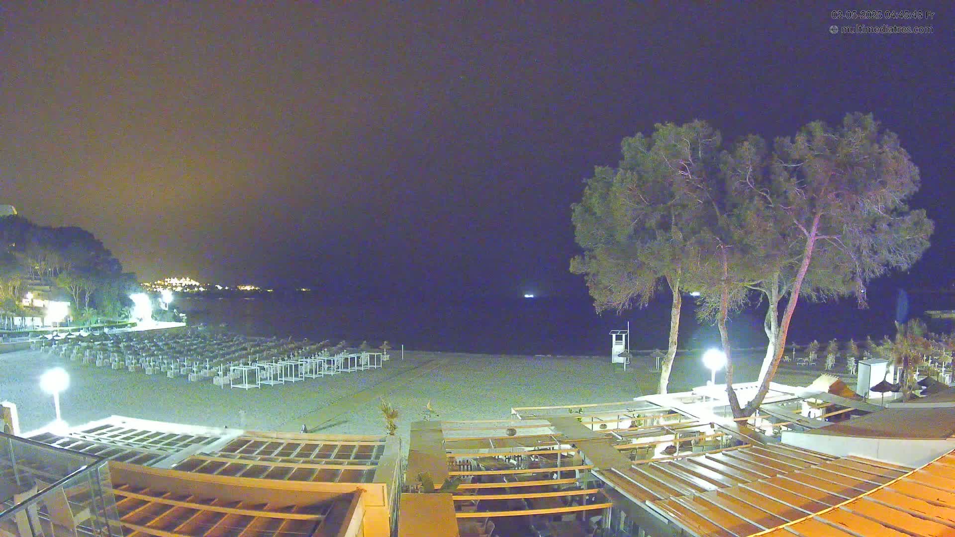 A nighttime view of a beach with rows of closed beach chairs and umbrellas, a few trees, and an outdoor restaurant area under a clear, dark sky.