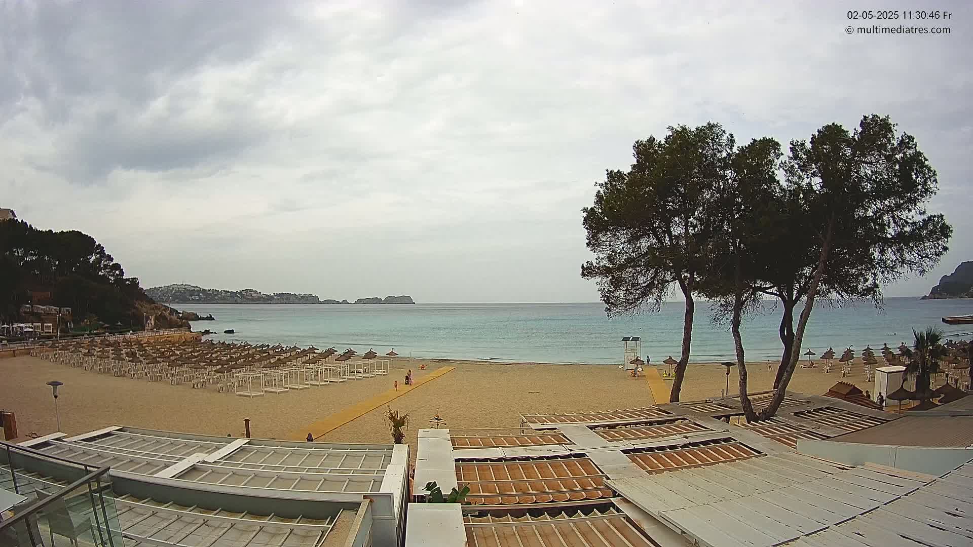 A mostly sunny beach with rows of beach umbrellas and a calm, light blue sea under a cloudy sky is viewed from atop a building.
