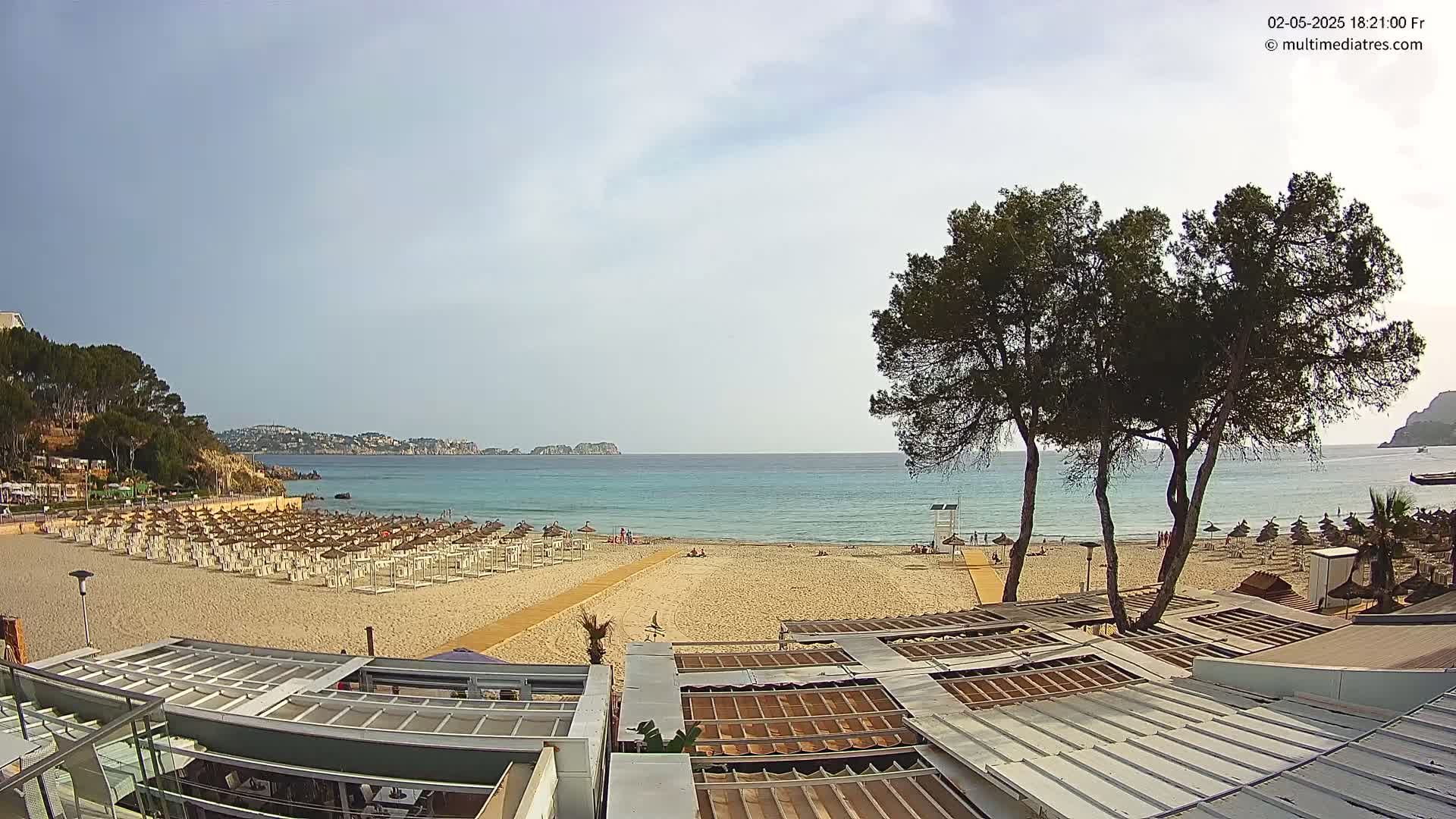 A sandy beach with numerous white umbrellas and a calm, turquoise sea under a mostly cloudy sky is viewed from a rooftop vantage point.