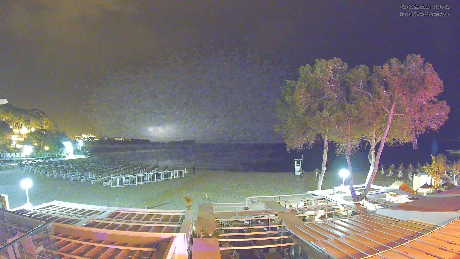 A nighttime view of a beach with rows of closed beach umbrellas, several trees, and a building with a patio overlooking the calm sea under a cloudy sky.