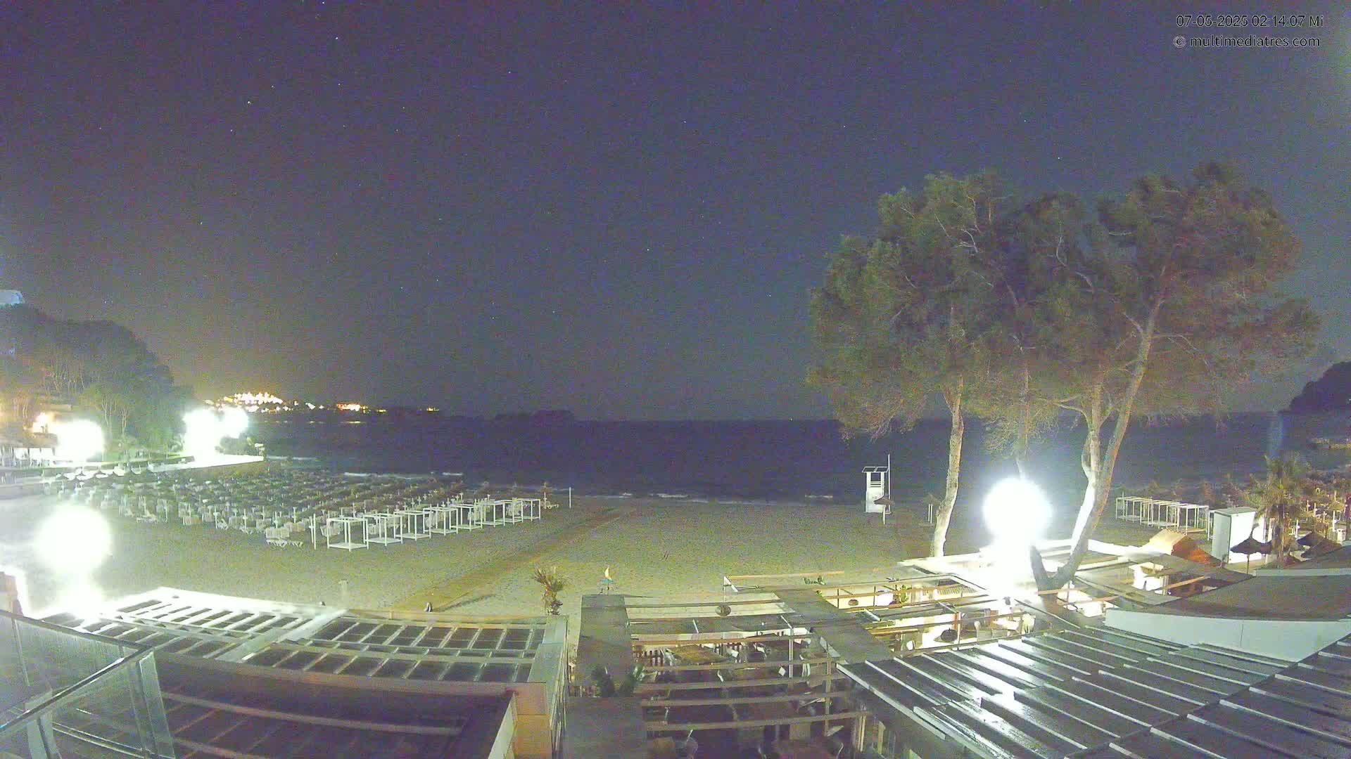 A mostly empty beach at night under a starry sky, with rows of beach cabanas, trees, and the lights of buildings visible.