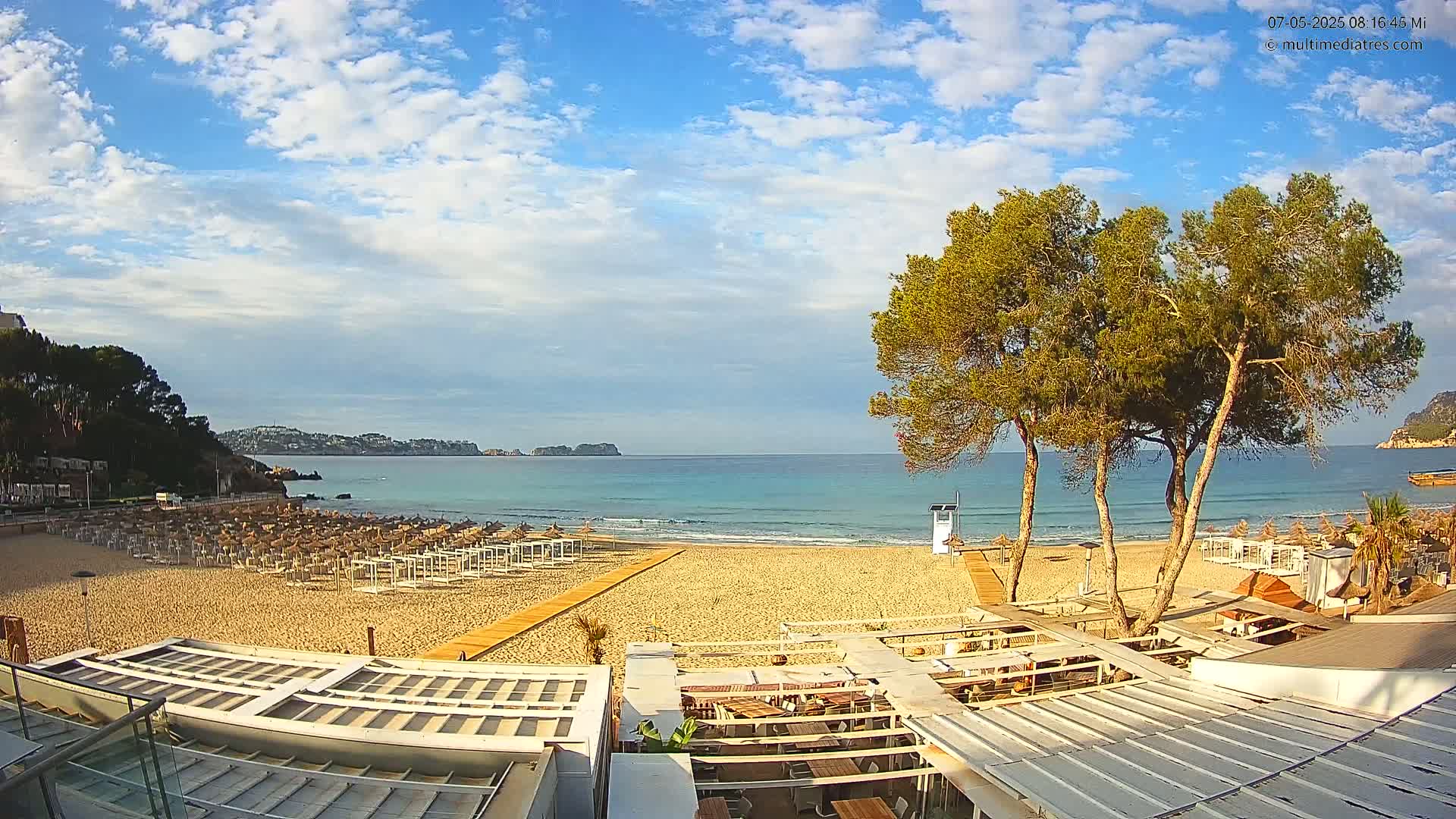 A sandy beach with rows of beach umbrellas and a beachfront restaurant is shown under a partly cloudy sky, overlooking a calm, turquoise ocean.