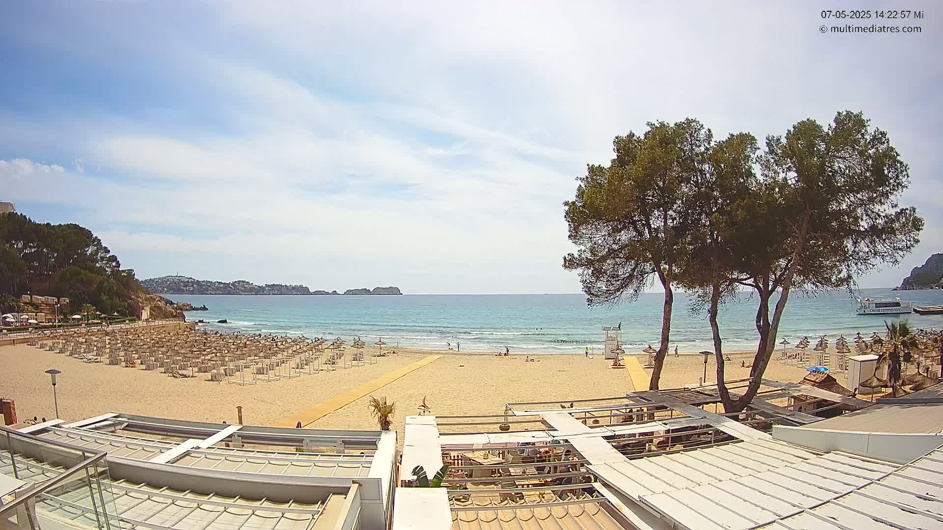 A sandy beach with numerous umbrellas and a few people is visible, overlooking a calm, blue ocean under a partly cloudy sky; tall pine trees frame the view.