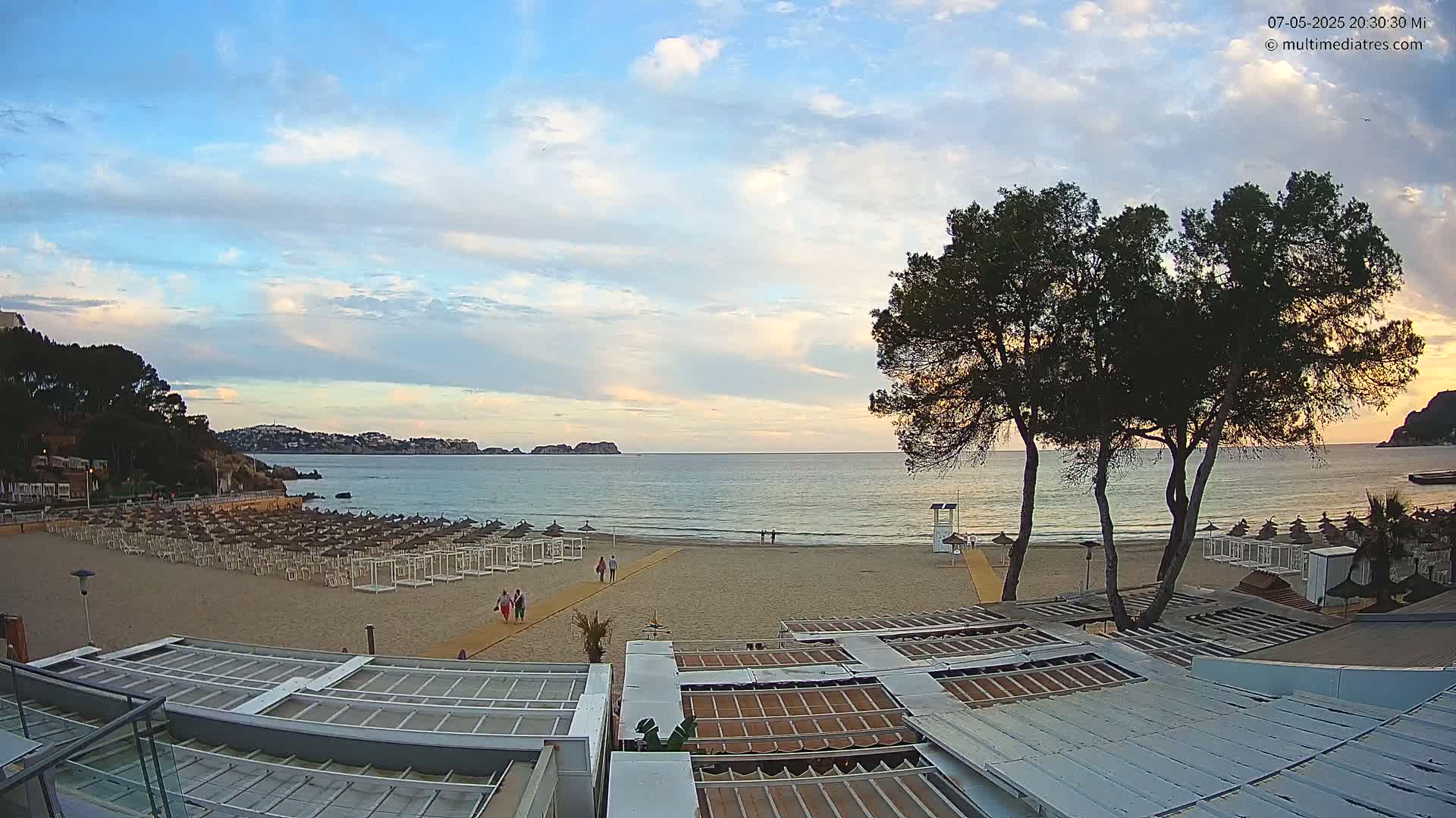 A sandy beach with numerous closed umbrellas and a few people walking on it, under a partly cloudy sunset sky, overlooks a calm sea with hills in the distance.