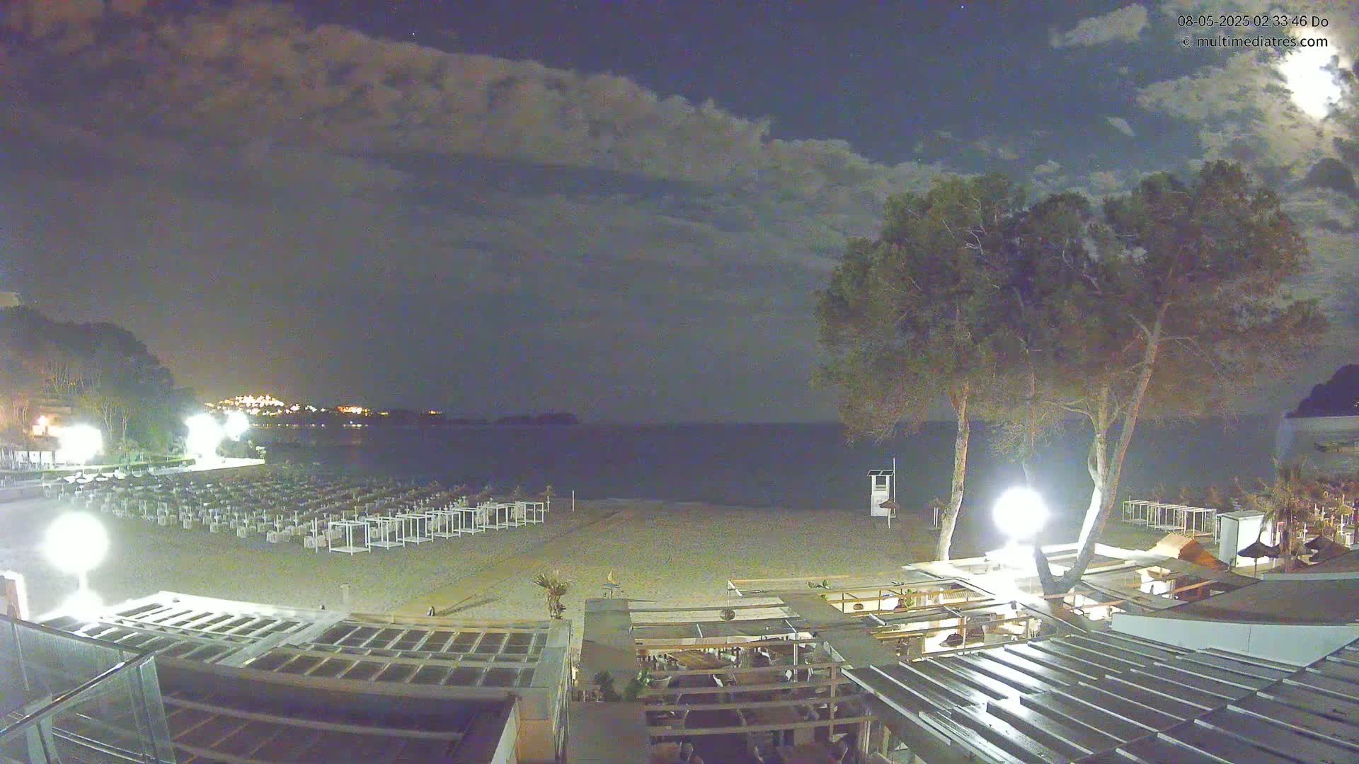 A nighttime beach scene under a partly cloudy sky shows rows of beach chairs on a sandy beach next to a calm sea, with trees and outdoor lighting illuminating a beachside restaurant.