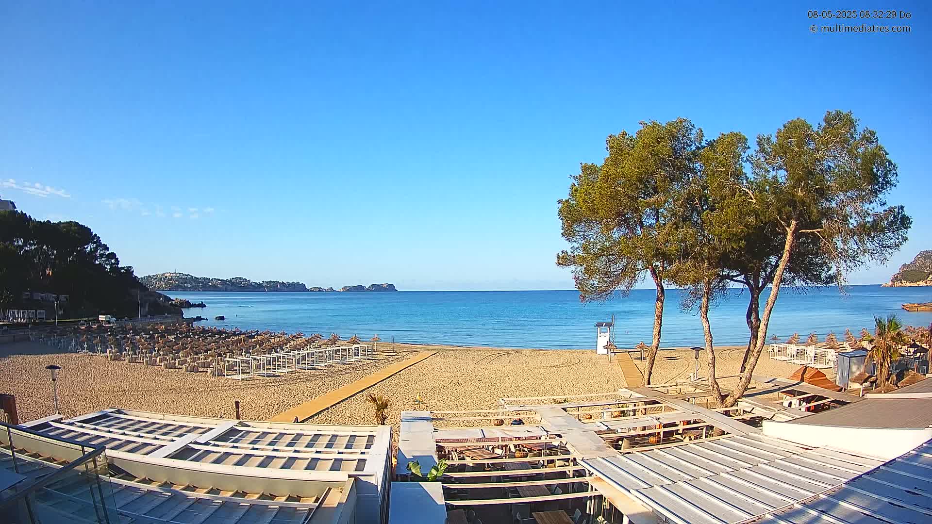 A sandy beach with rows of closed beach umbrellas, several trees, and a lifeguard stand is visible under a clear blue sky, overlooking a calm sea with distant hills.