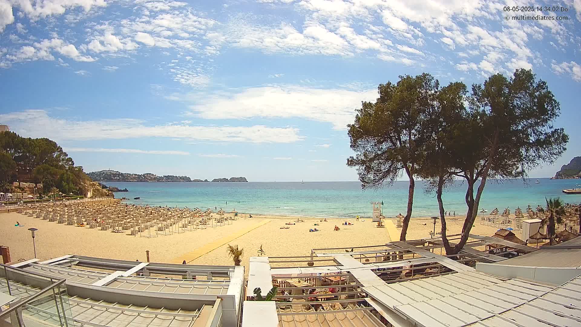 A sandy beach with numerous umbrellas and a few people is situated next to a calm, turquoise ocean under a partly cloudy blue sky.