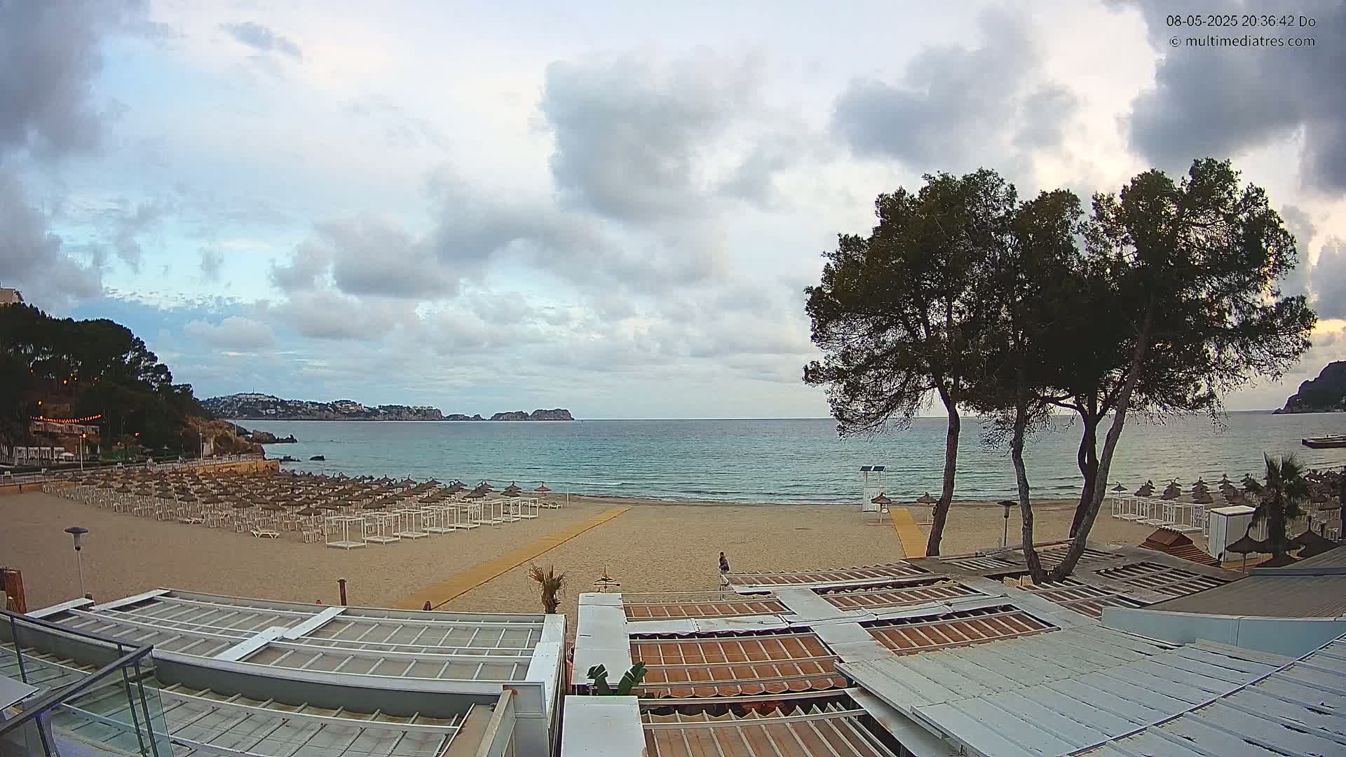 A sandy beach with rows of beach umbrellas and a calm sea under a partly cloudy sky is viewed from above a beachfront building.