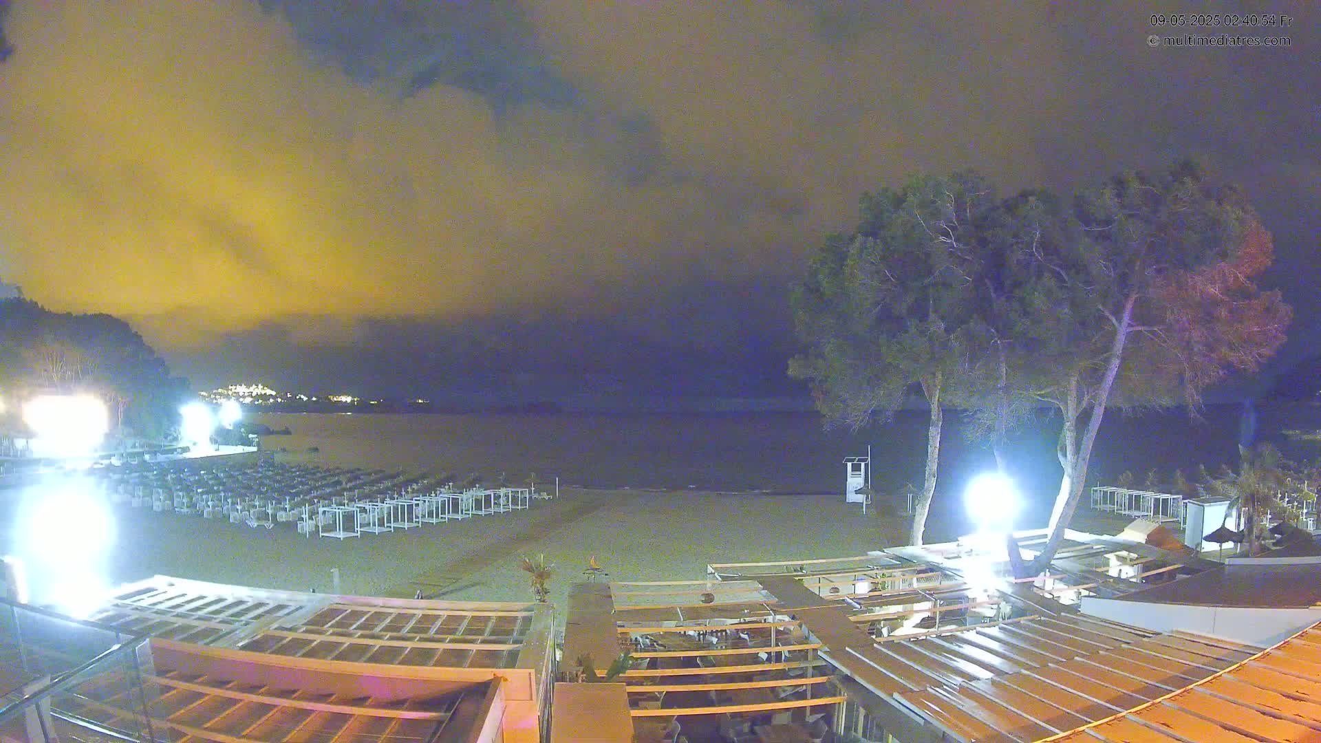 An empty beach at night under a cloudy, yellowish sky is shown, with rows of closed beach umbrellas and lounge chairs on the sand, illuminated by lights near some trees and buildings.