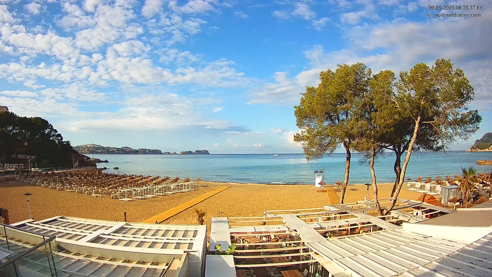 A sandy beach with rows of beach umbrellas and a beachfront restaurant is seen under a mostly sunny sky with scattered clouds, overlooking a calm sea with distant hills.