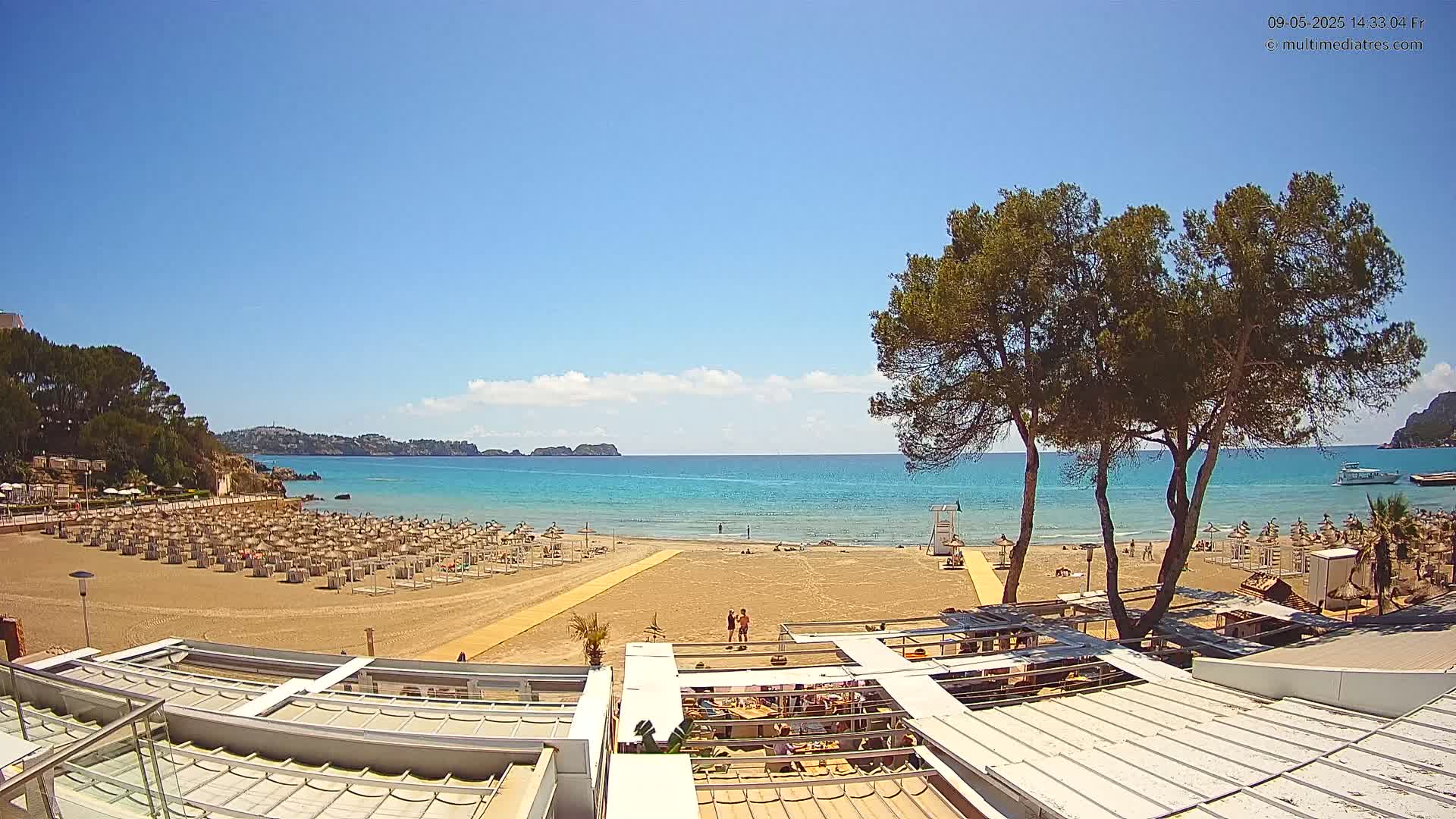 A sandy beach with numerous umbrellas and a few people is visible under a clear blue sky, overlooking a calm turquoise sea with distant hills.