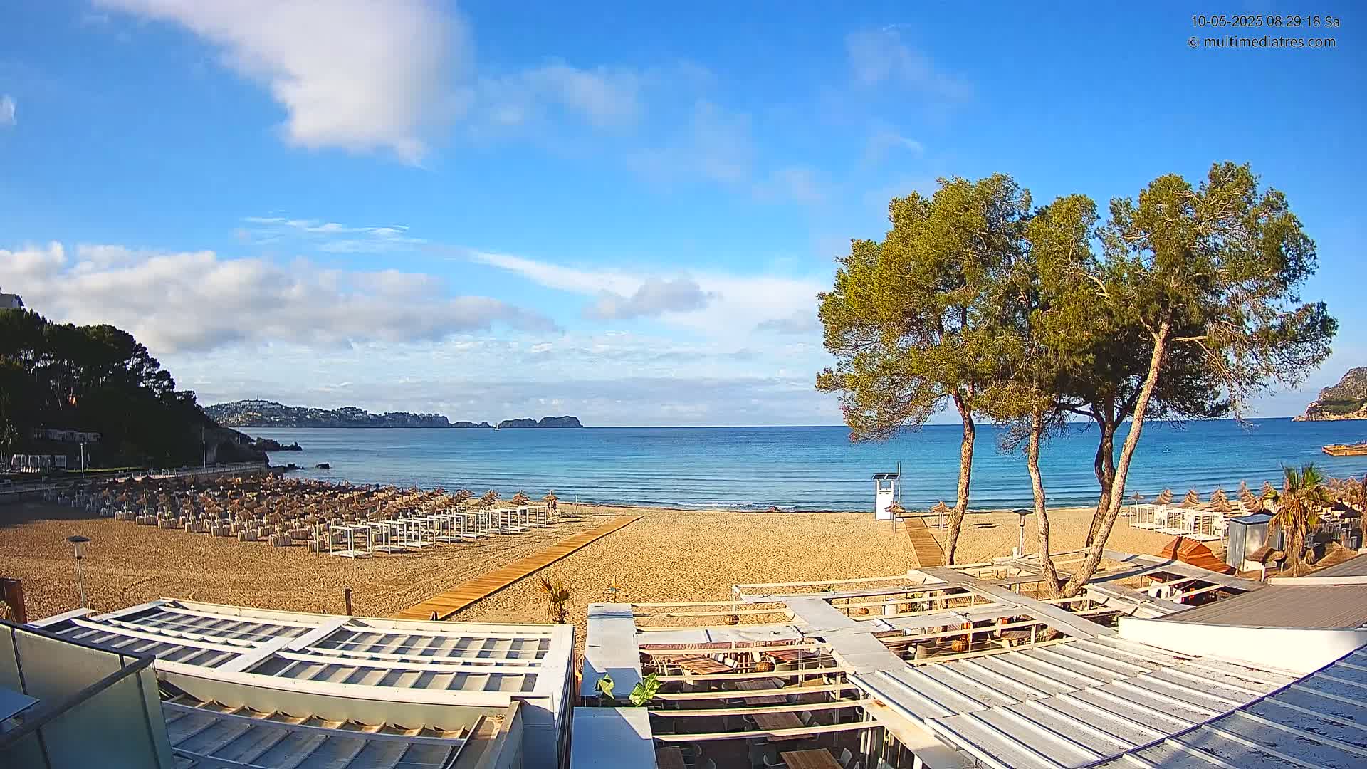 A sandy beach with rows of beach umbrellas and lounge chairs stretches to a calm, blue sea under a mostly sunny sky with some scattered clouds.
