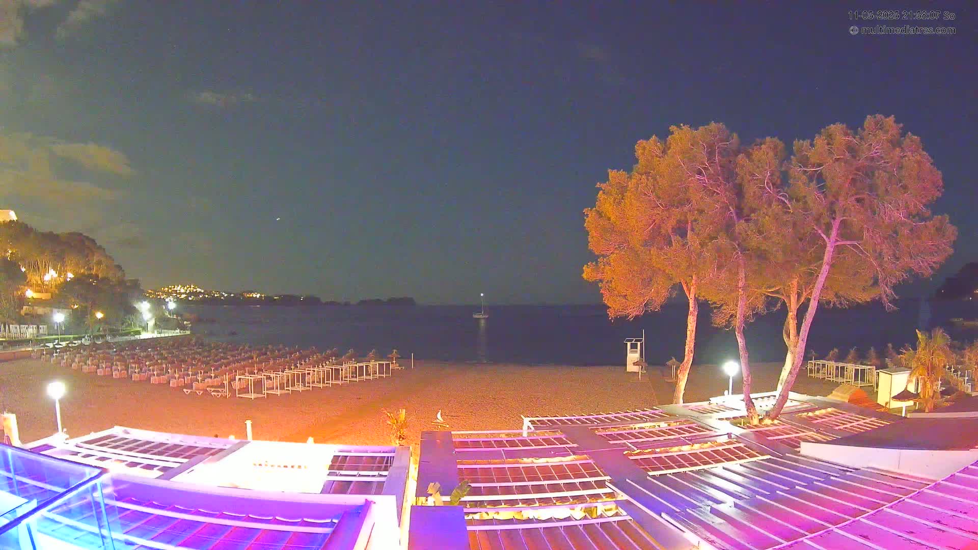 A nighttime beach scene shows a sandy shore with rows of beach furniture, illuminated trees, and a calm body of water with a single sailboat in the distance under a mostly clear sky.