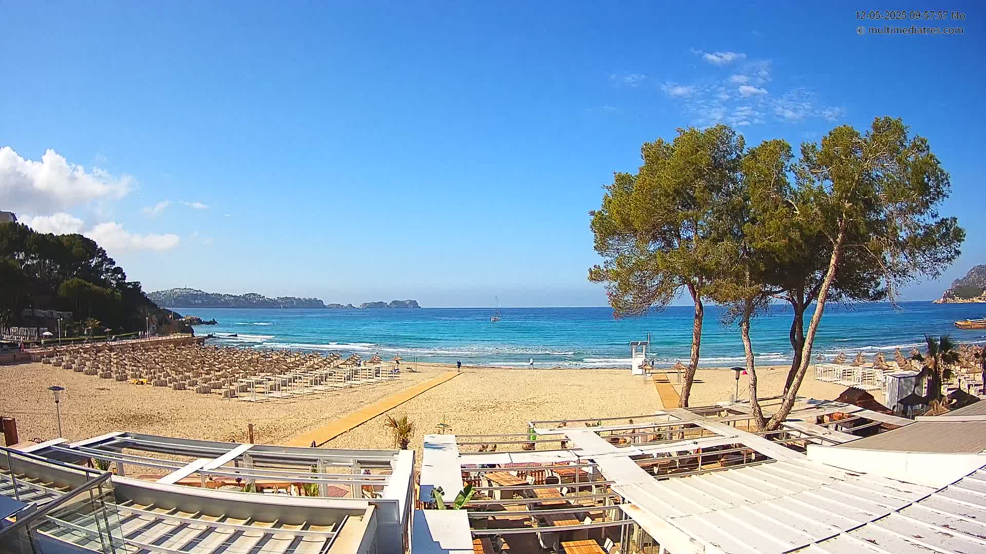 A sandy beach with rows of beach umbrellas, a boardwalk, and a beachfront restaurant is visible under a clear blue sky, with calm turquoise water and a few small boats in the distance.