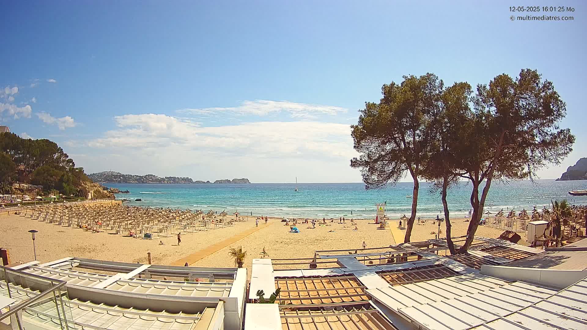 A sandy beach with numerous umbrellas and sunbathers is visible, overlooking a calm, turquoise ocean under a mostly sunny sky with a few scattered clouds.
