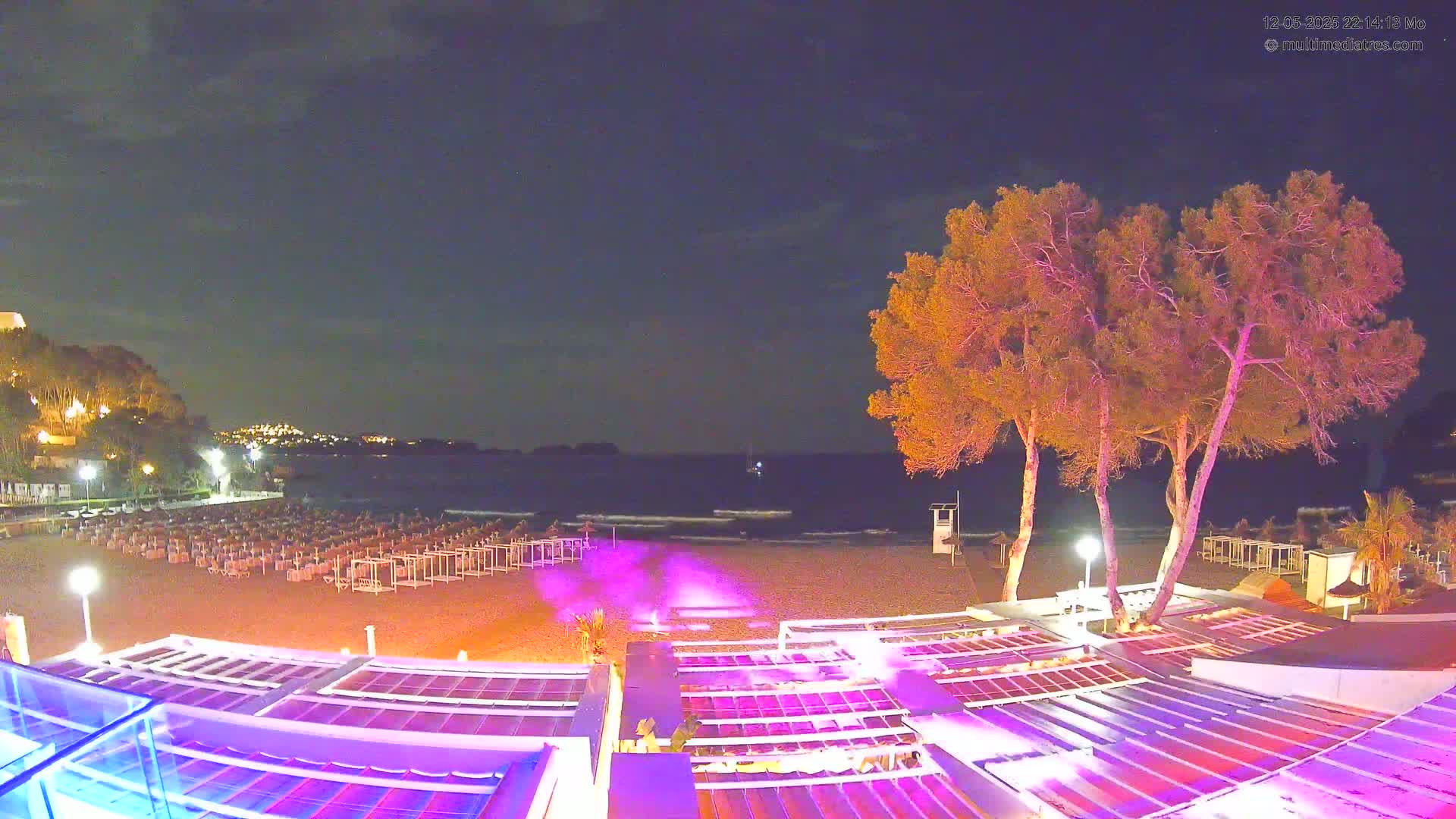 A nighttime beach scene shows a sandy shore with rows of beach chairs, illuminated trees, and a building with a brightly lit roof overlooking a calm sea under a dark sky.