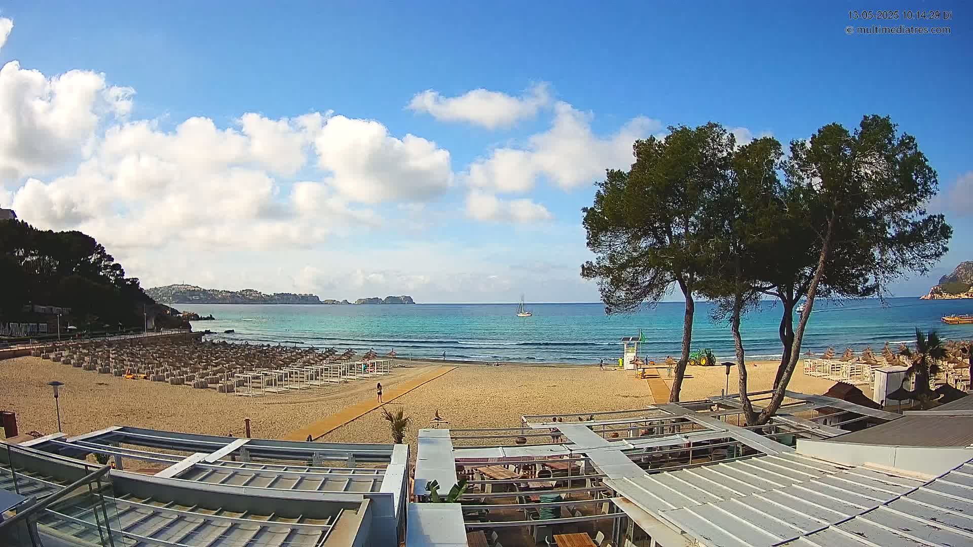 A mostly sunny day at a beach with calm, turquoise water, rows of beach umbrellas, and several trees near a beachfront building.