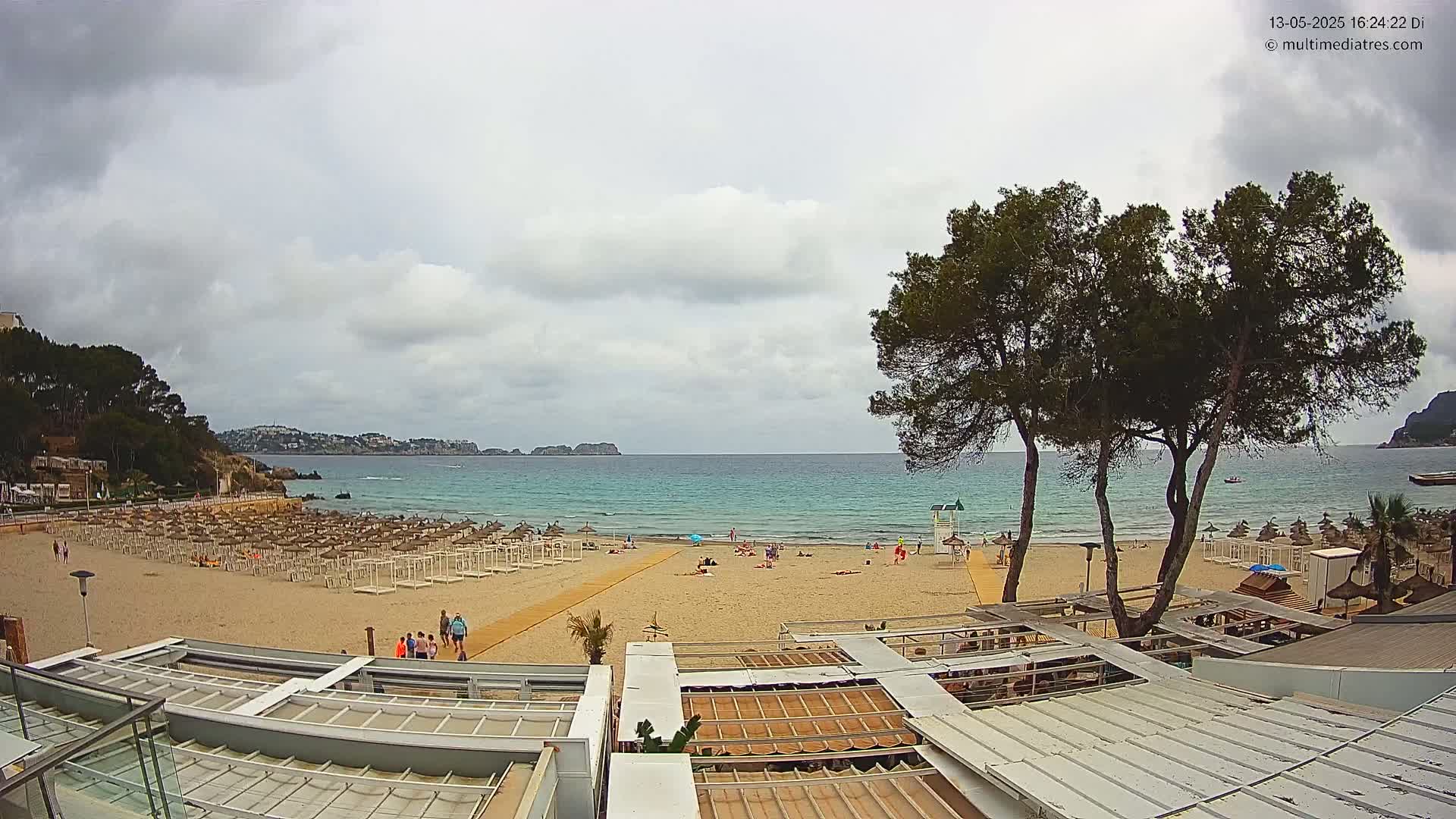A mostly cloudy day overlooks a sandy beach with scattered beachgoers and umbrellas, a calm turquoise sea, and dark green trees in the foreground.