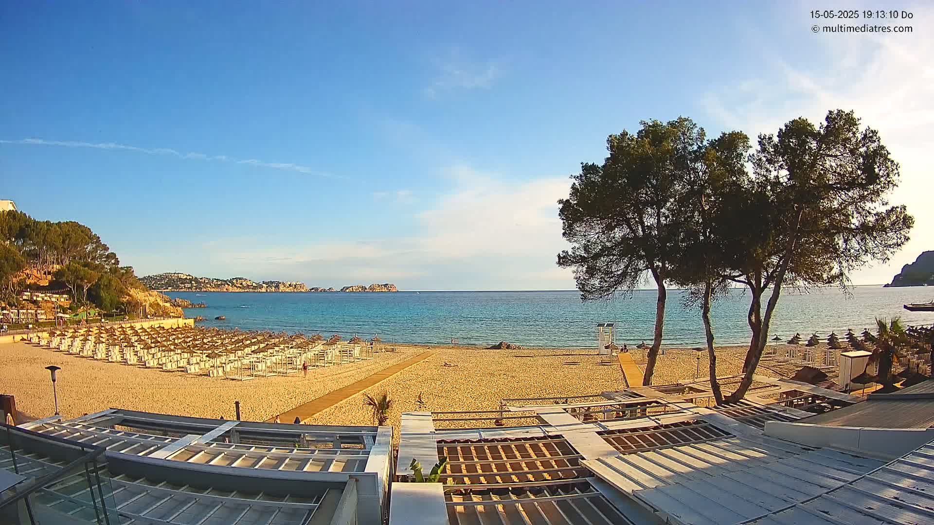 A sandy beach with rows of beach umbrellas overlooks a calm, turquoise ocean under a mostly clear, sunny sky.