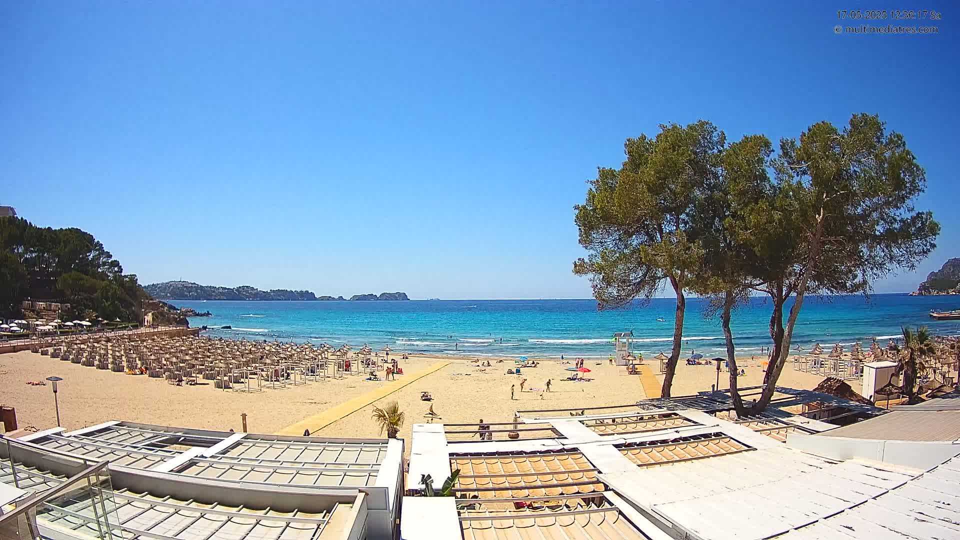 A sandy beach with numerous umbrellas and a few people on it, overlooks a calm, turquoise ocean under a clear blue sky.