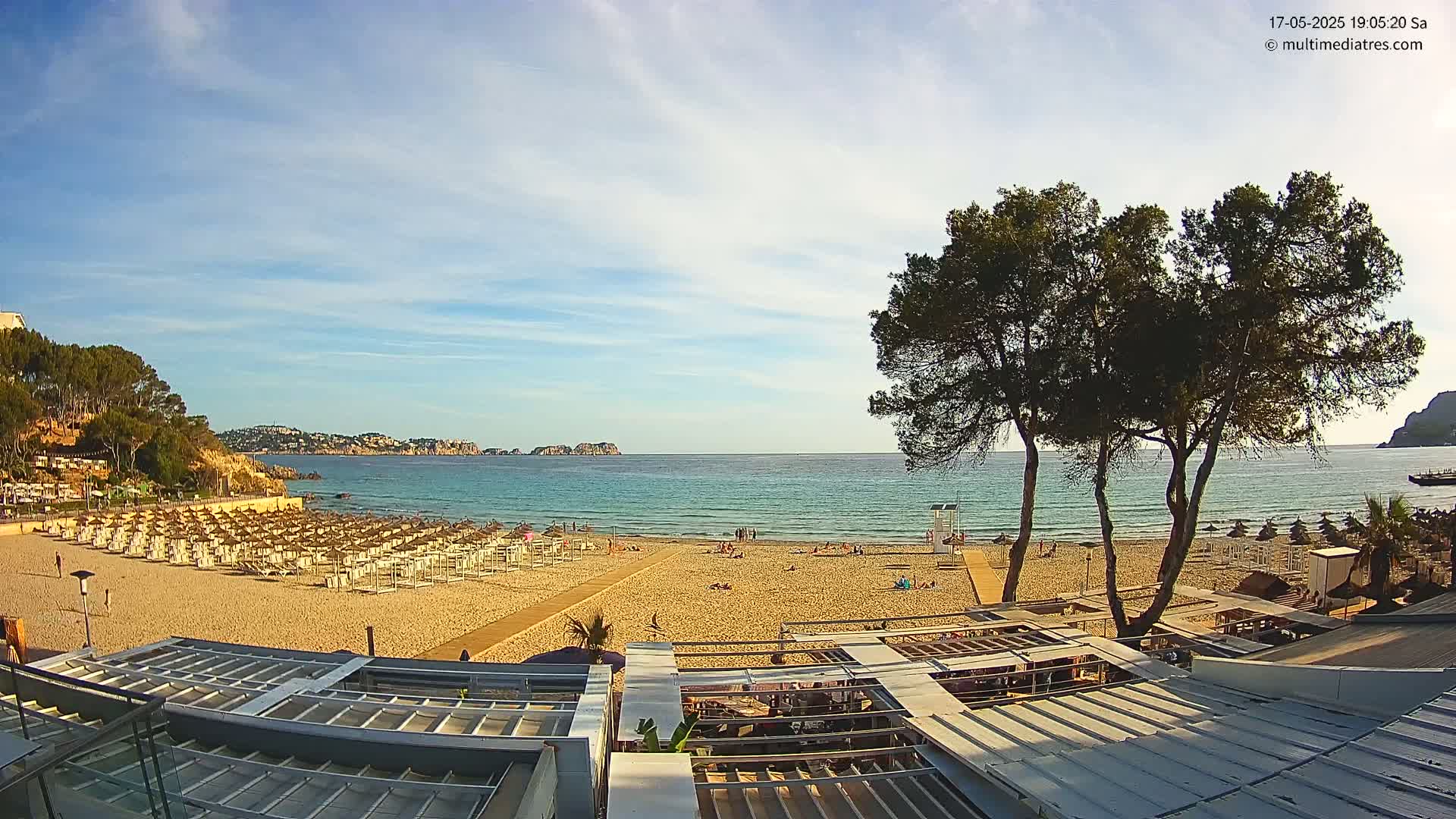 A sandy beach with numerous beach umbrellas and a few people on it, overlooks a calm, turquoise sea under a mostly sunny sky.