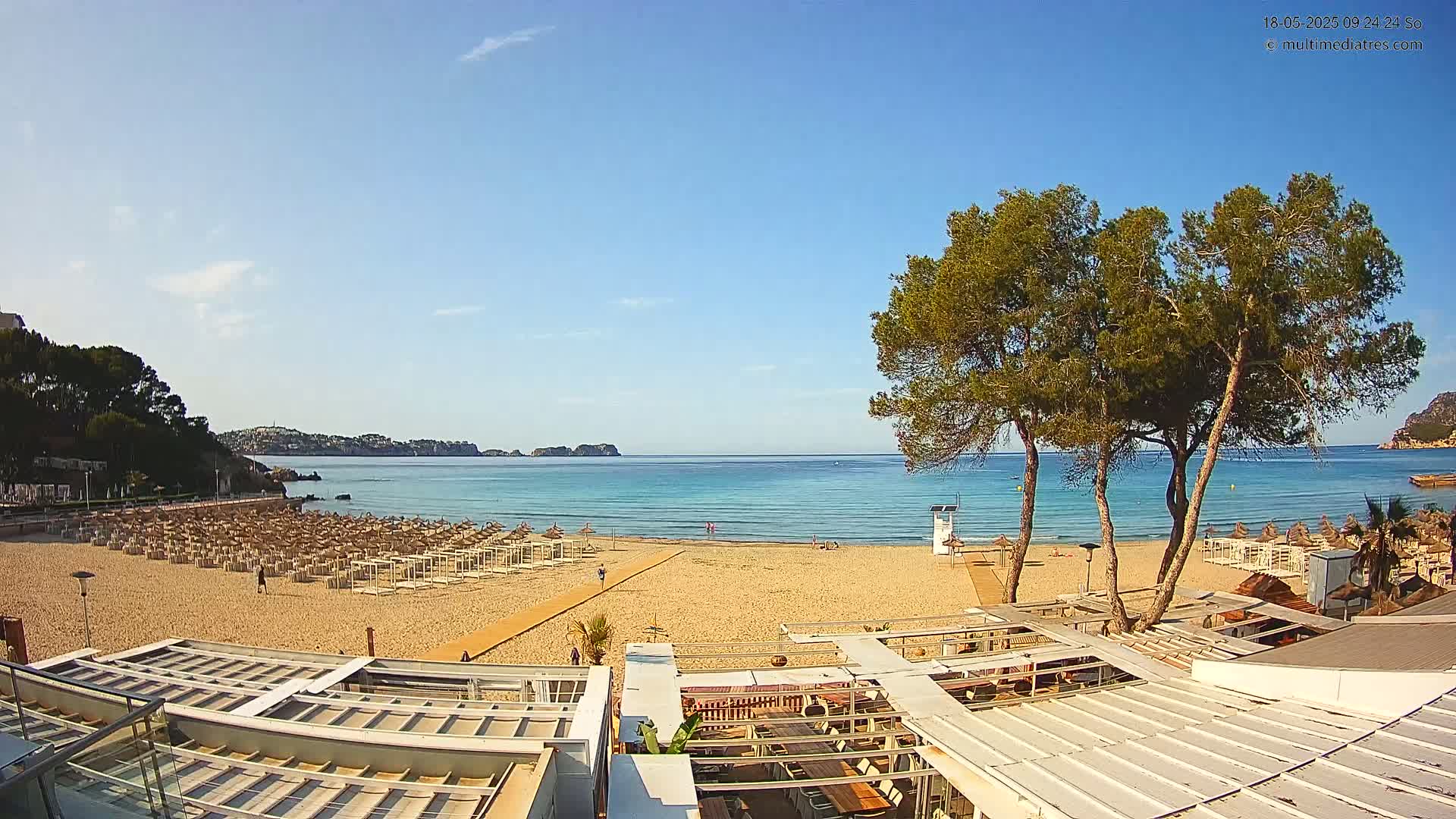 A sandy beach with rows of beach umbrellas and a calm, clear ocean under a sunny sky is seen from above a beachside restaurant.