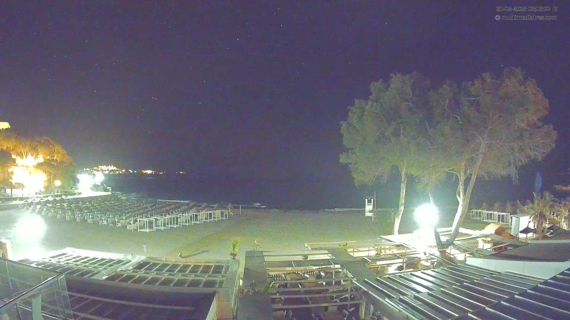 A nighttime beach scene shows rows of beach chairs on the sand, a few trees, a building, and a calm sea under a clear, starry sky.