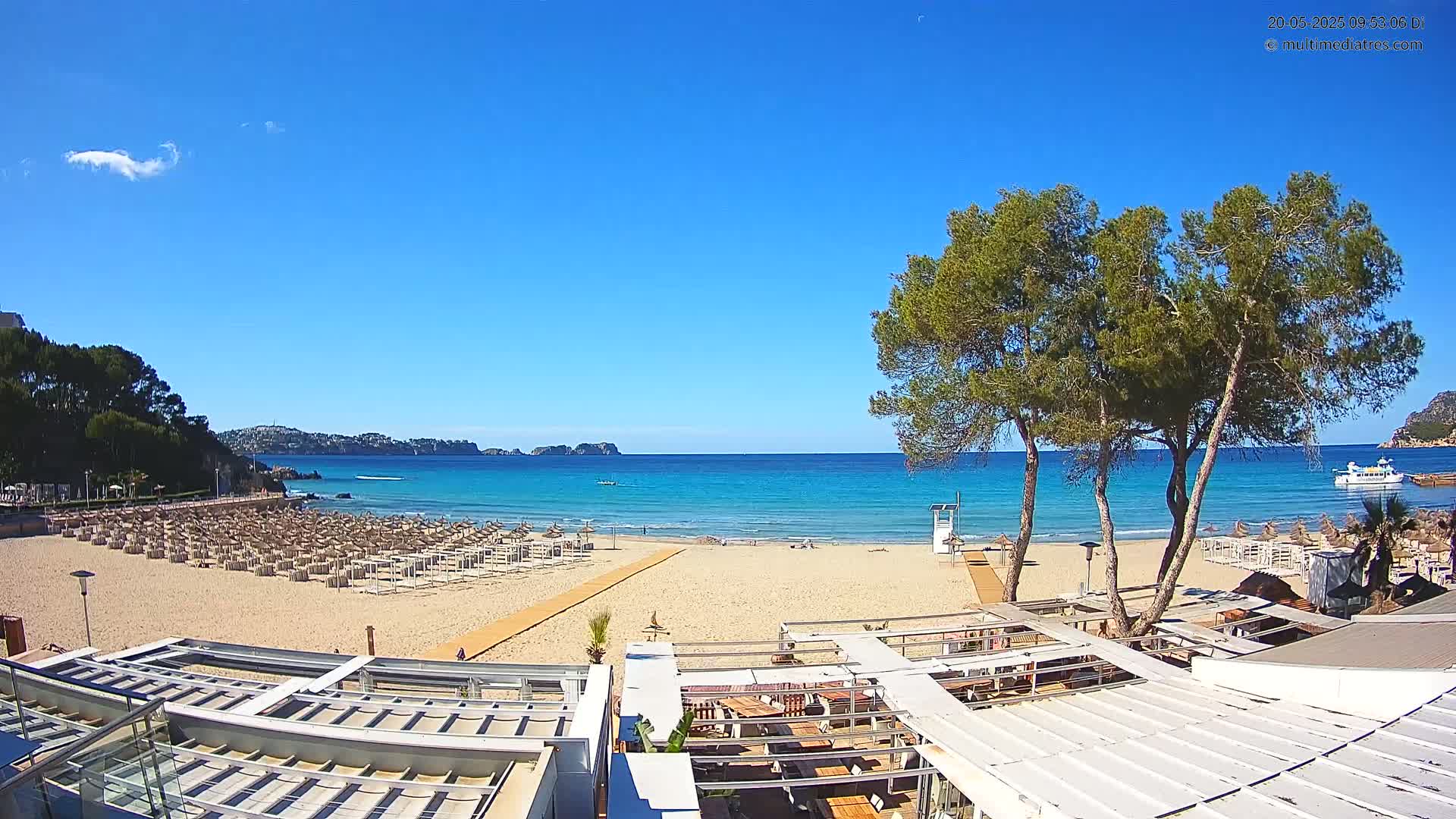 A sandy beach with numerous lounge chairs, a boardwalk, and a beachfront restaurant is visible, all under a clear blue sky.