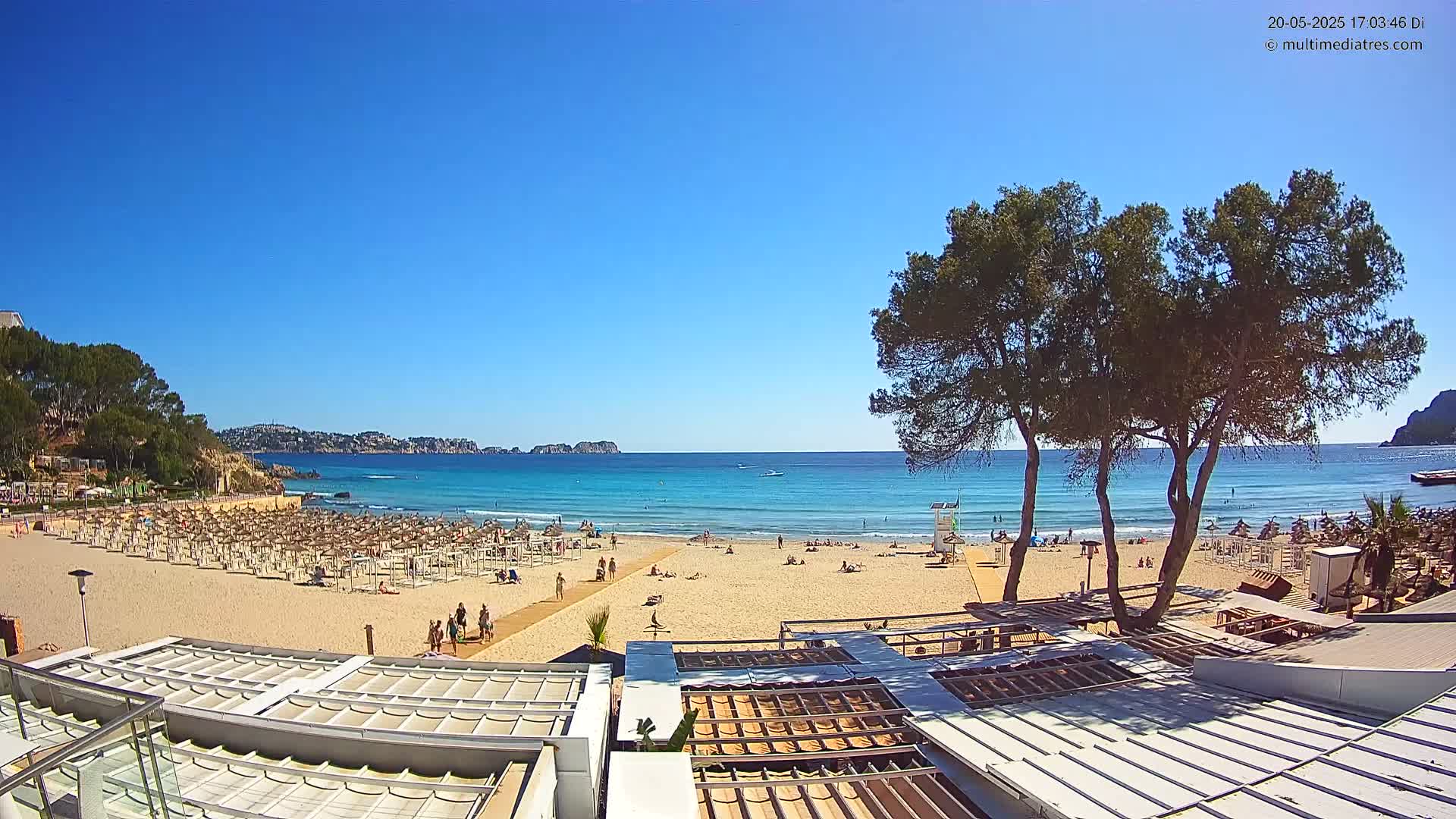 A sandy beach with numerous beach umbrellas and sunbathers stretches towards a calm, turquoise ocean under a clear blue sky.