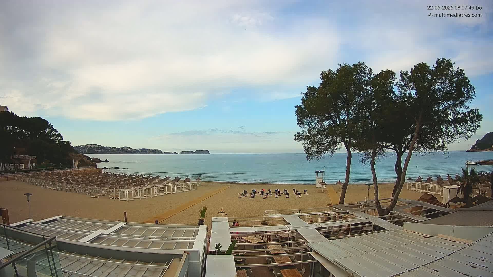 A sandy beach with rows of beach umbrellas and a group of people exercising outdoors under a partly cloudy sky, overlooks a calm sea with distant hills.