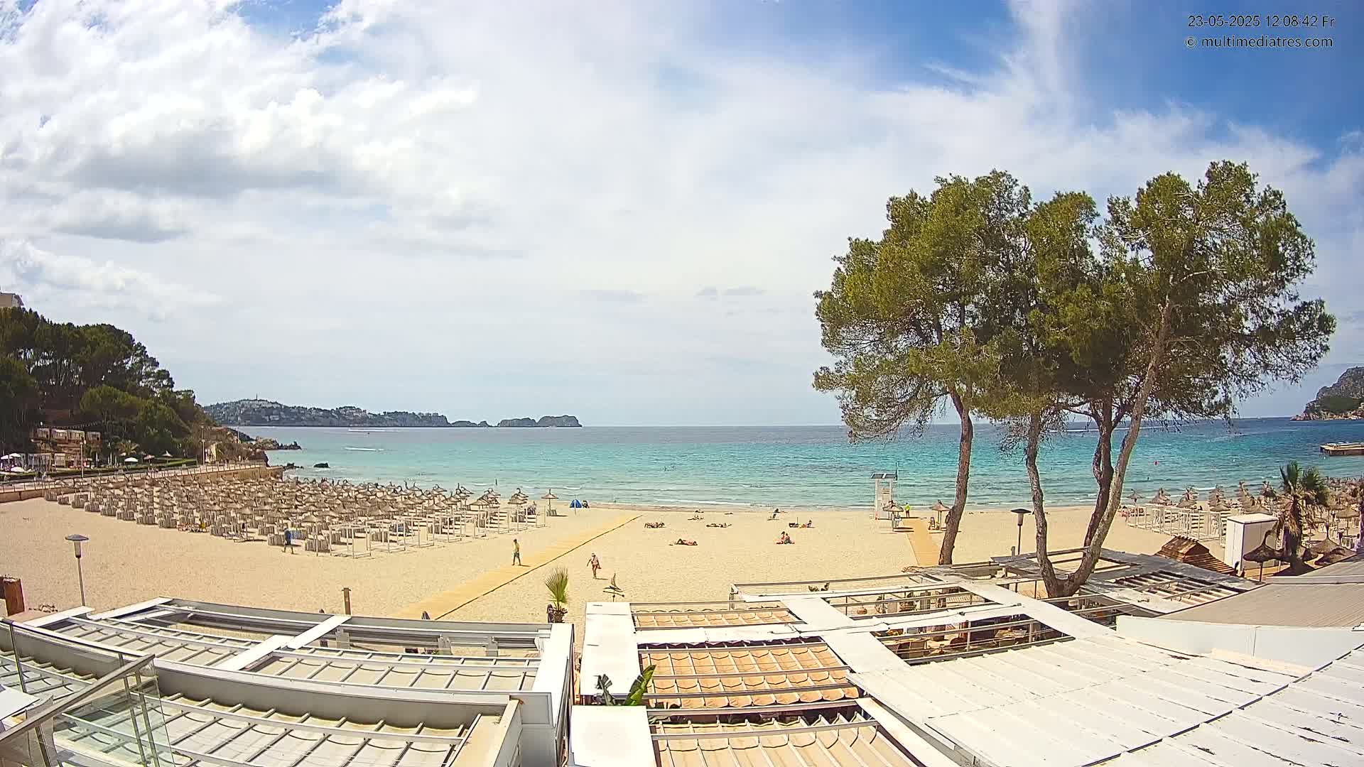 A sandy beach with numerous lounge chairs and umbrellas is visible under a partly cloudy sky, with calm turquoise water and a distant shoreline in the background.
