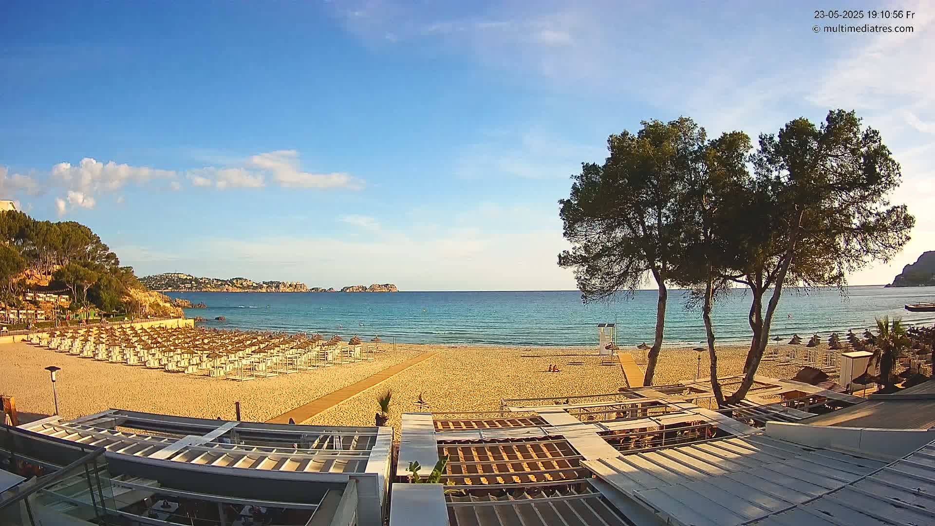 A sandy beach with rows of beach umbrellas and a calm, clear ocean under a mostly sunny sky is viewed from a rooftop terrace.