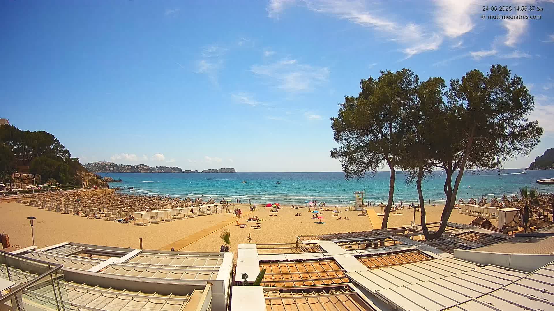 A sunny beach scene shows a sandy shore with numerous beach umbrellas and lounge chairs, several people relaxing, and a calm turquoise sea under a mostly clear blue sky.