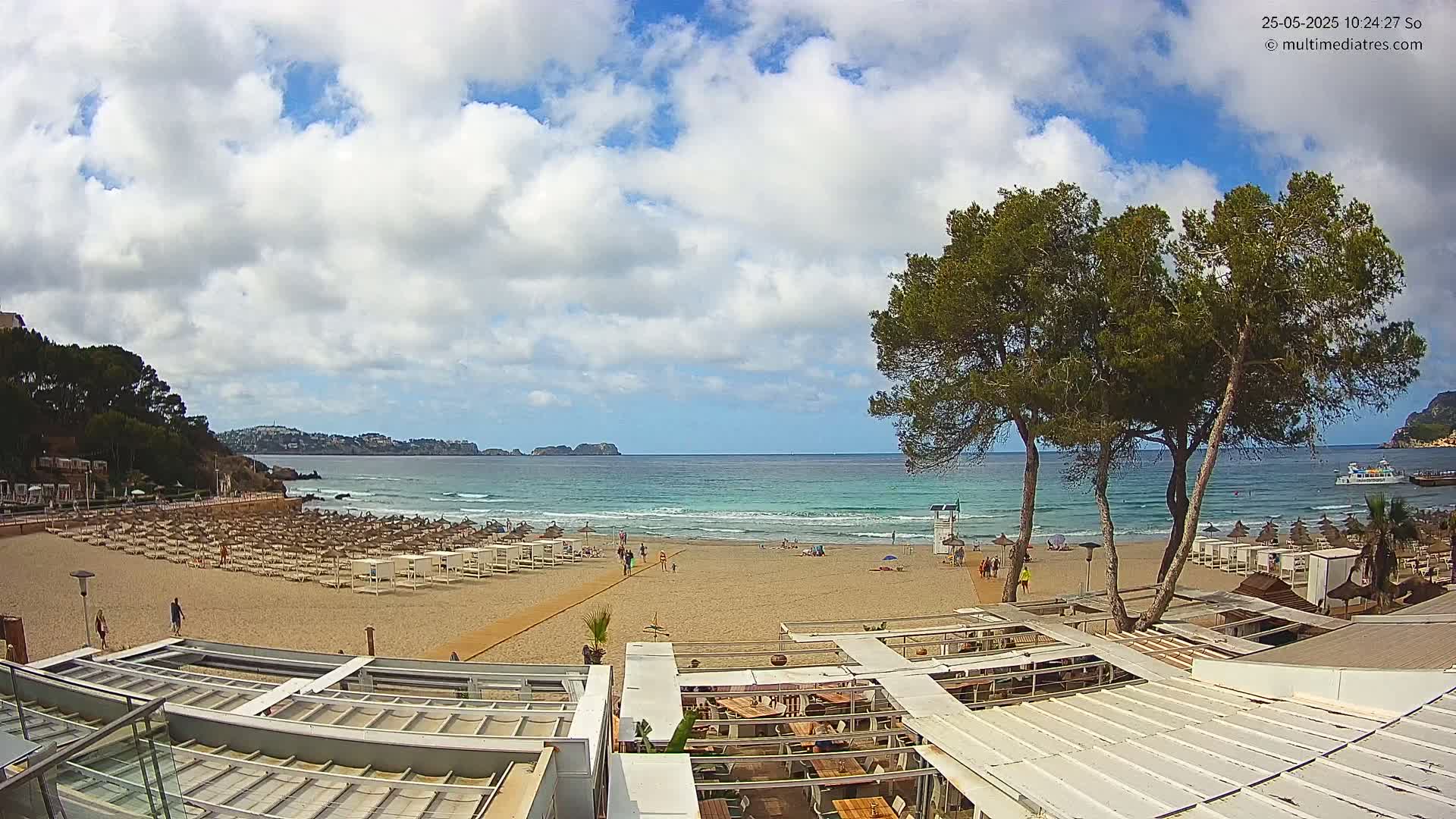 A partly cloudy day at a sandy beach with calm ocean waves, beach umbrellas, and several people present, viewed from a partially covered outdoor patio.
