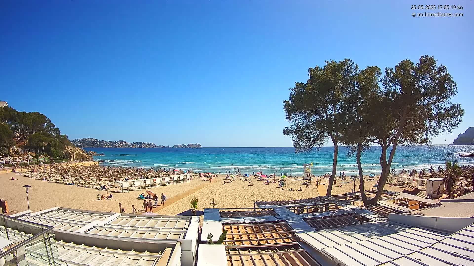 A sunny beach scene shows a sandy shore with numerous beach umbrellas and sunbathers, calm turquoise water, and several trees in the foreground.