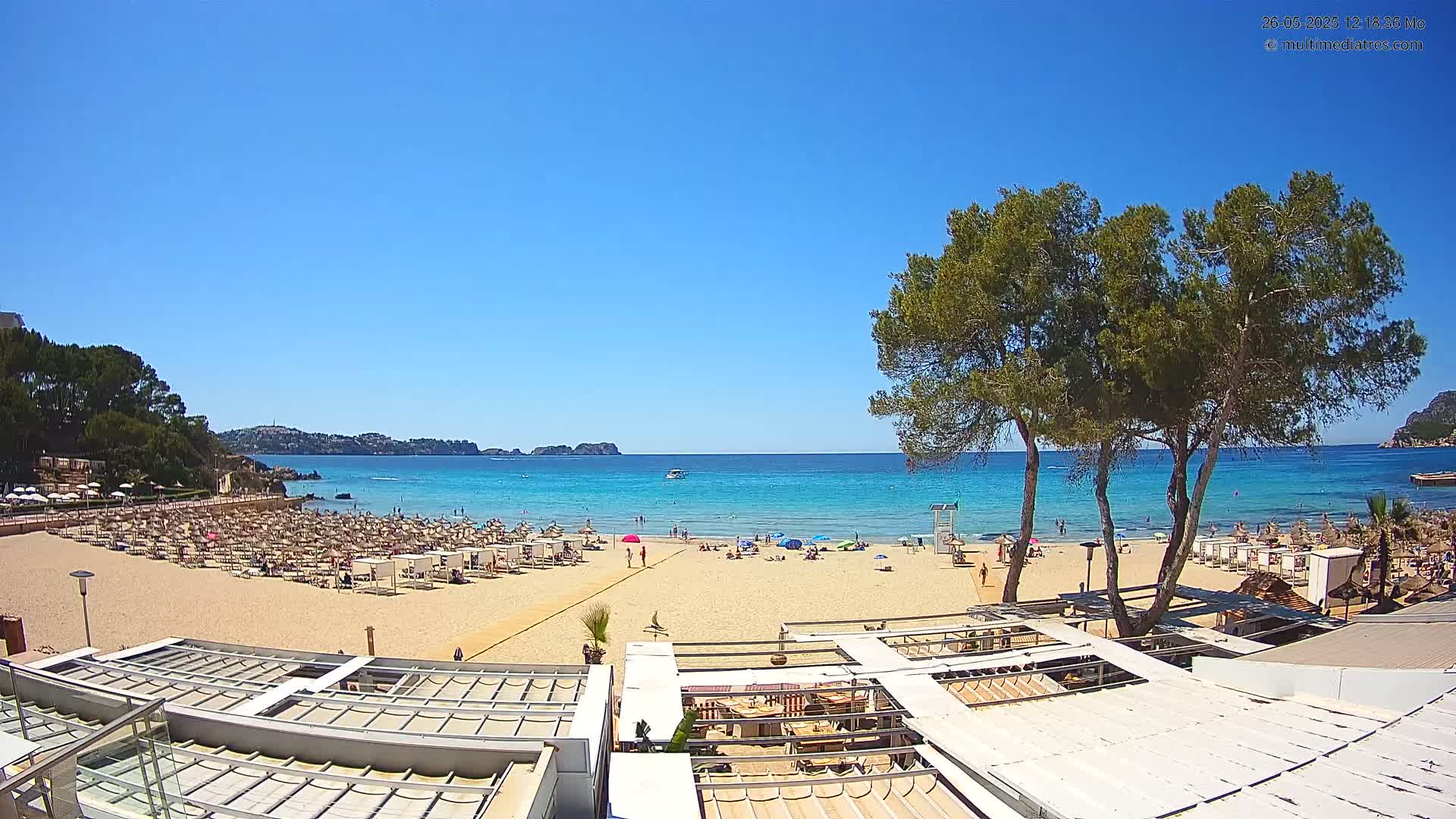 A sandy beach with numerous umbrellas and beachgoers is visible, overlooked by a beachfront restaurant under a clear blue sky.