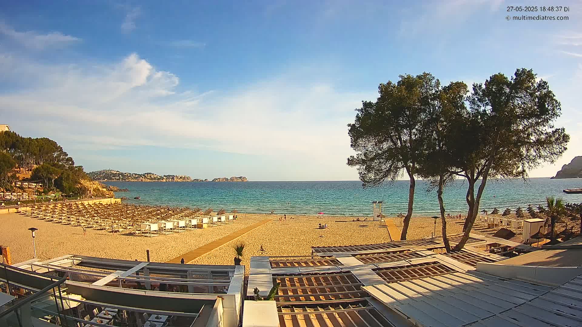 A sandy beach with numerous beach umbrellas and a calm, clear turquoise ocean under a mostly sunny sky is viewed from atop a beachside building.