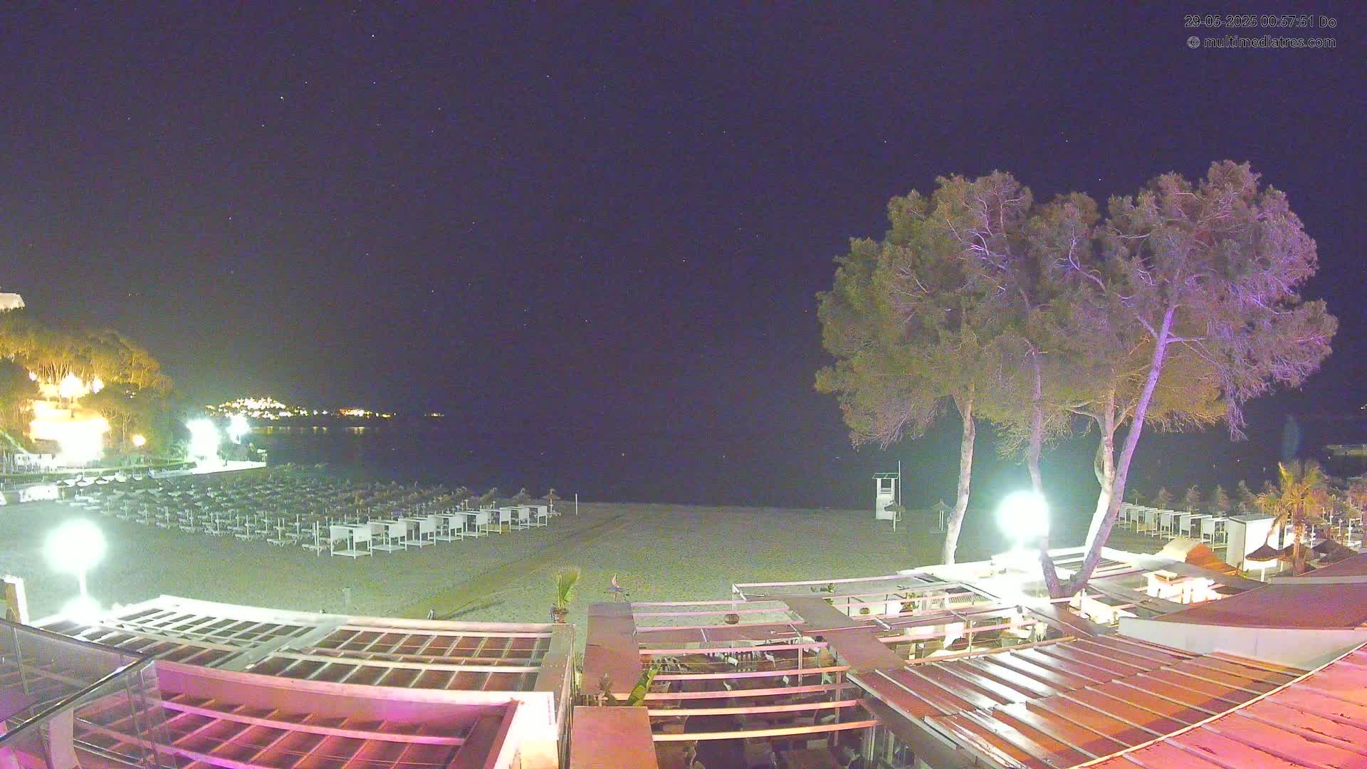 A nighttime view of a mostly empty beach with rows of closed beach chairs, a few trees, and a building with outdoor seating under a clear, starry sky.