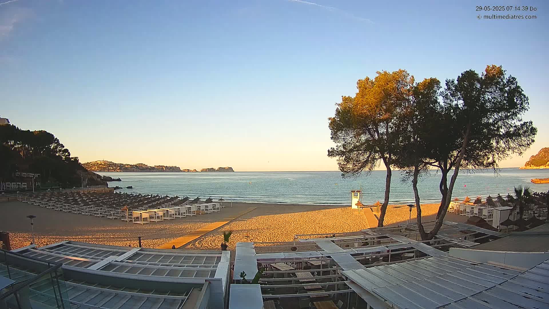 A sandy beach with rows of closed beach umbrellas and a lifeguard stand is shown at sunrise, overlooking calm, clear water and hills in the distance.