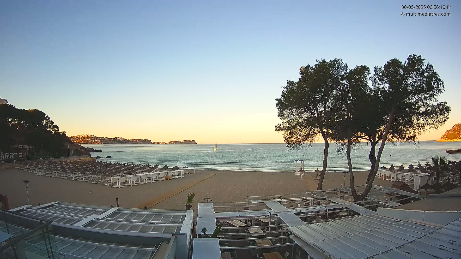 A sandy beach with rows of lounge chairs is visible at sunrise, with calm ocean water and hills in the background.