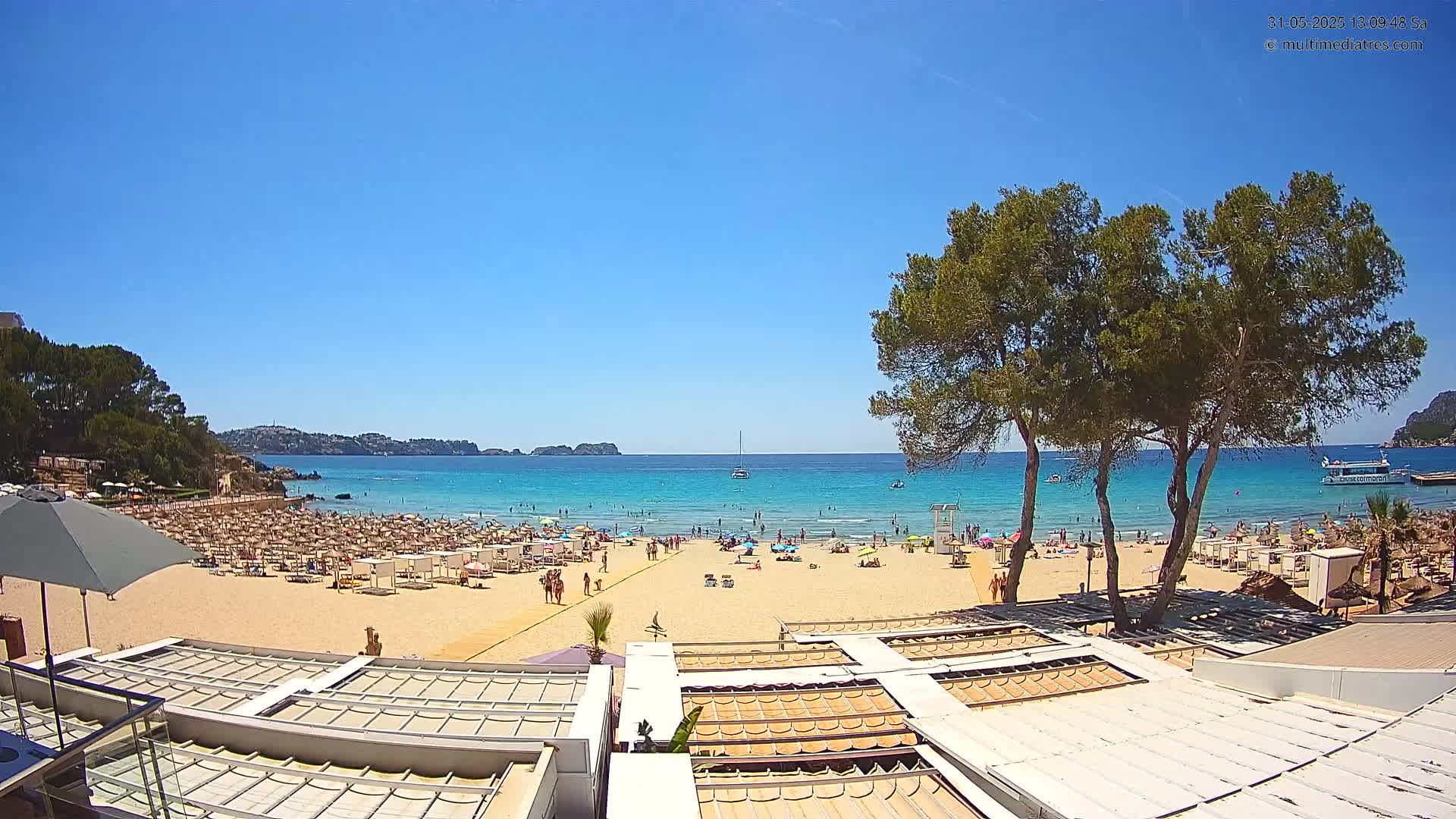 A sandy beach with numerous sunbathers and beach umbrellas under a clear blue sky, bordered by calm turquoise water and green vegetation.