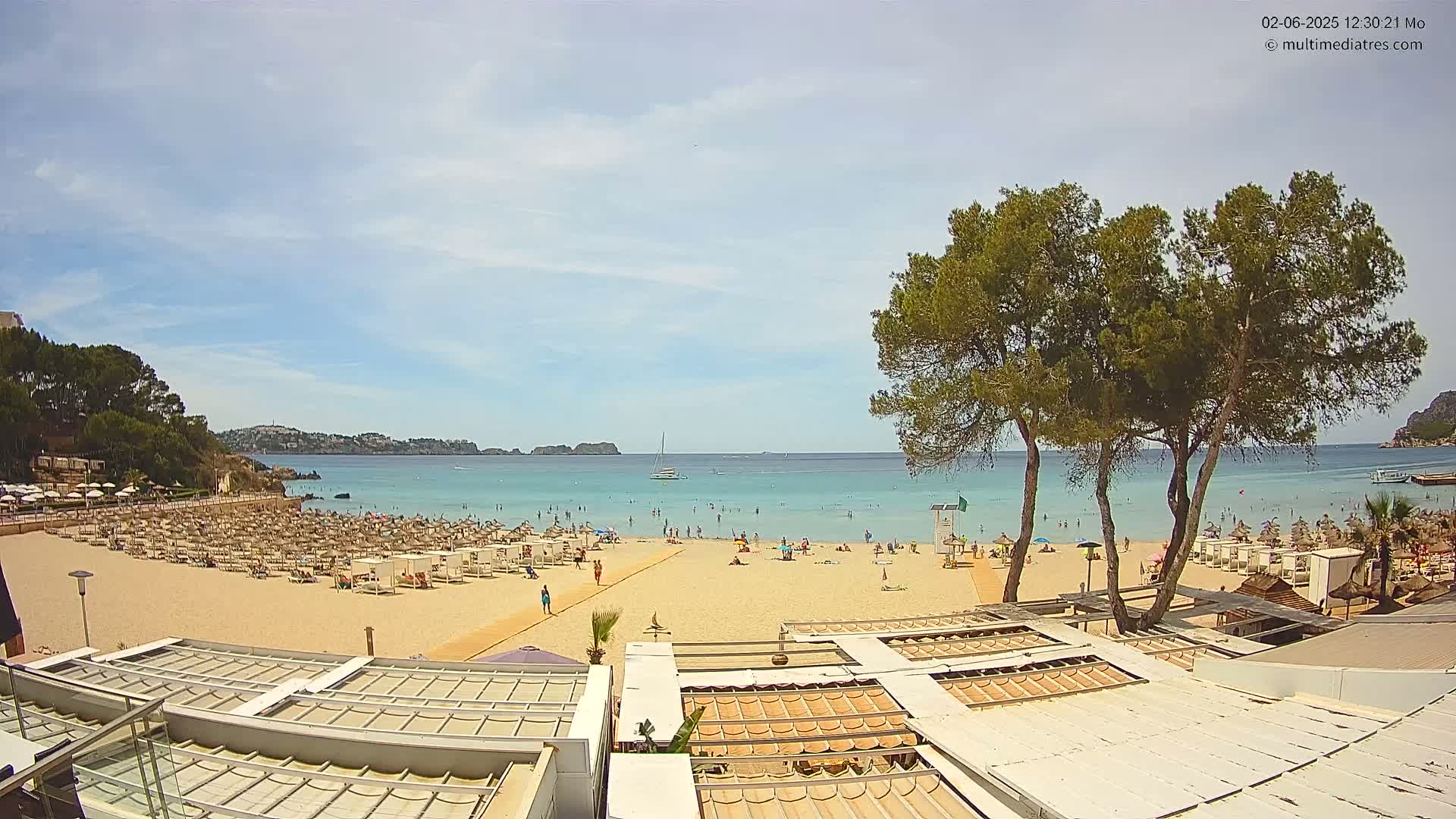A sandy beach with many beach umbrellas and people, backed by trees and buildings, is visible under a partly sunny sky on a clear day.