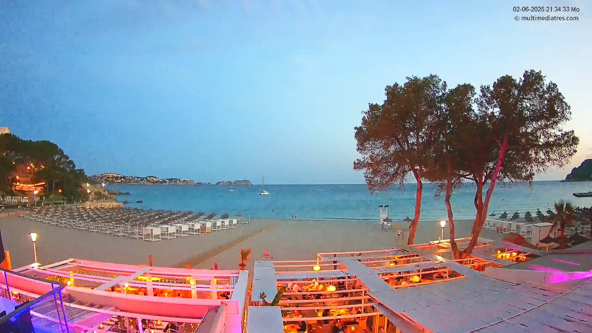A beach bar with pink lighting overlooks a calm, twilight ocean with a sandy beach lined with lounge chairs.