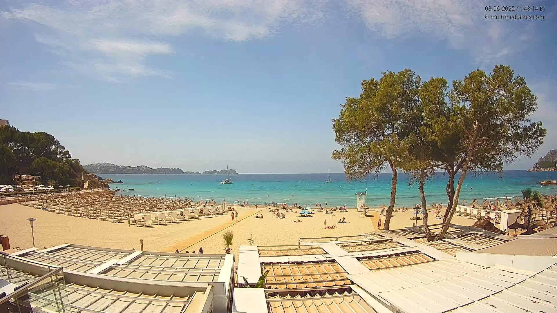 A sandy beach with many lounge chairs and umbrellas under a sunny sky, next to a calm turquoise sea with a few boats and people swimming and sunbathing.