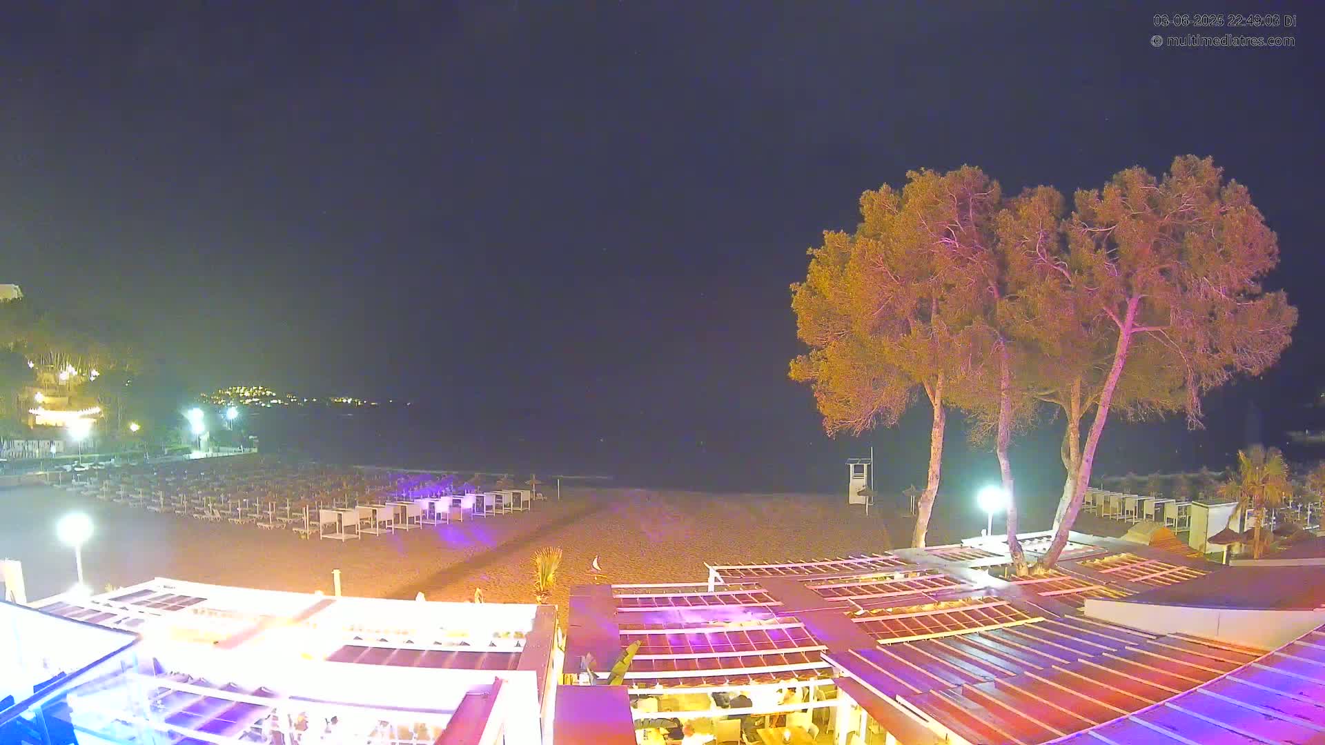 A nighttime view of a beach at night, showing rows of beach chairs and umbrellas, several trees near a building with illuminated outdoor seating, and a calm sea in the background.