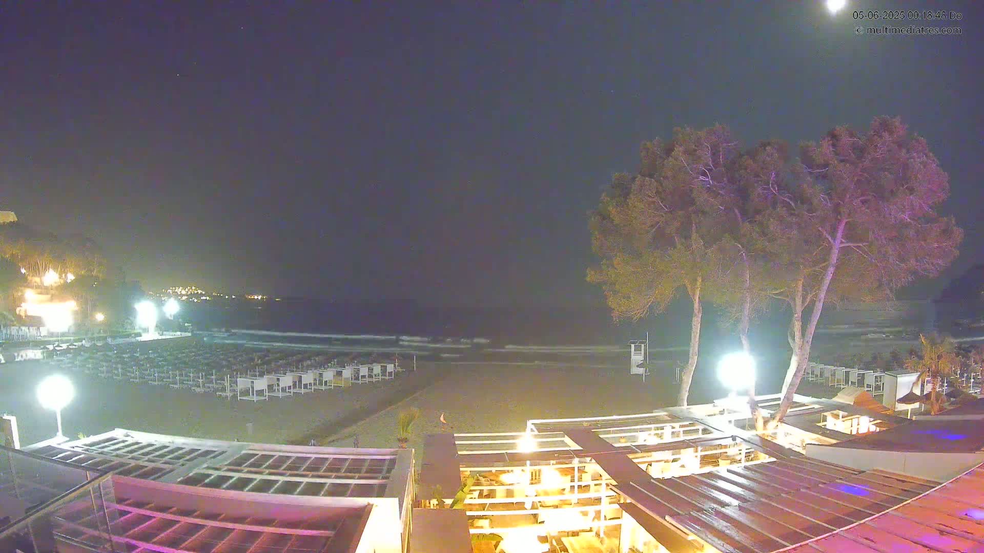 A nighttime beach scene shows gently waving water under a clear, dark sky, with rows of beach chairs and a beachfront restaurant illuminated by lights.